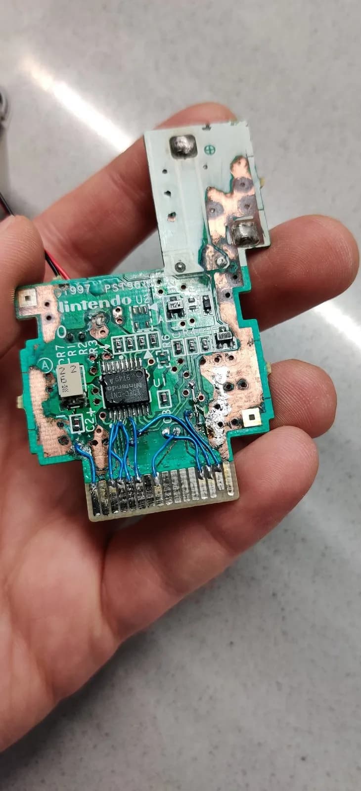 N64 Rumble Pak green circuit board after repair, held in a hand. A cluster of blue Kynar jumper wires bridges the broken traces and corroded vias left by the battery leak, the solder on every component has been touched up, and the copper now looks clean and shiny with most of the corrosion gone. The black motor-driver chip sits above.