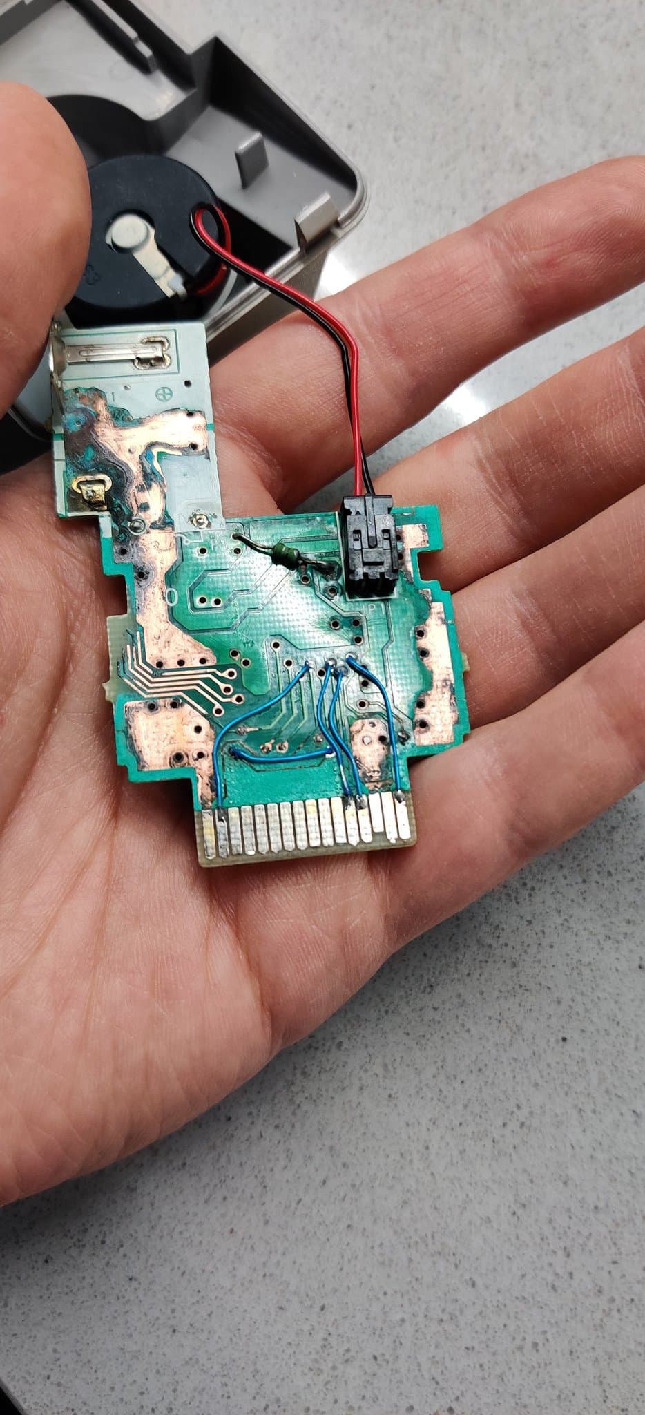 Disassembled N64 Rumble Pak green circuit board held in a hand after repair. Blue Kynar jumper wires bridge the broken traces and corroded vias, some running via to via and some down to the gold edge connector, following the board's layout to restore the connections. A small black connector carrying red and black wires is plugged into its receptacle on the board, and most of the battery corrosion has been cleaned away so the copper looks shiny.