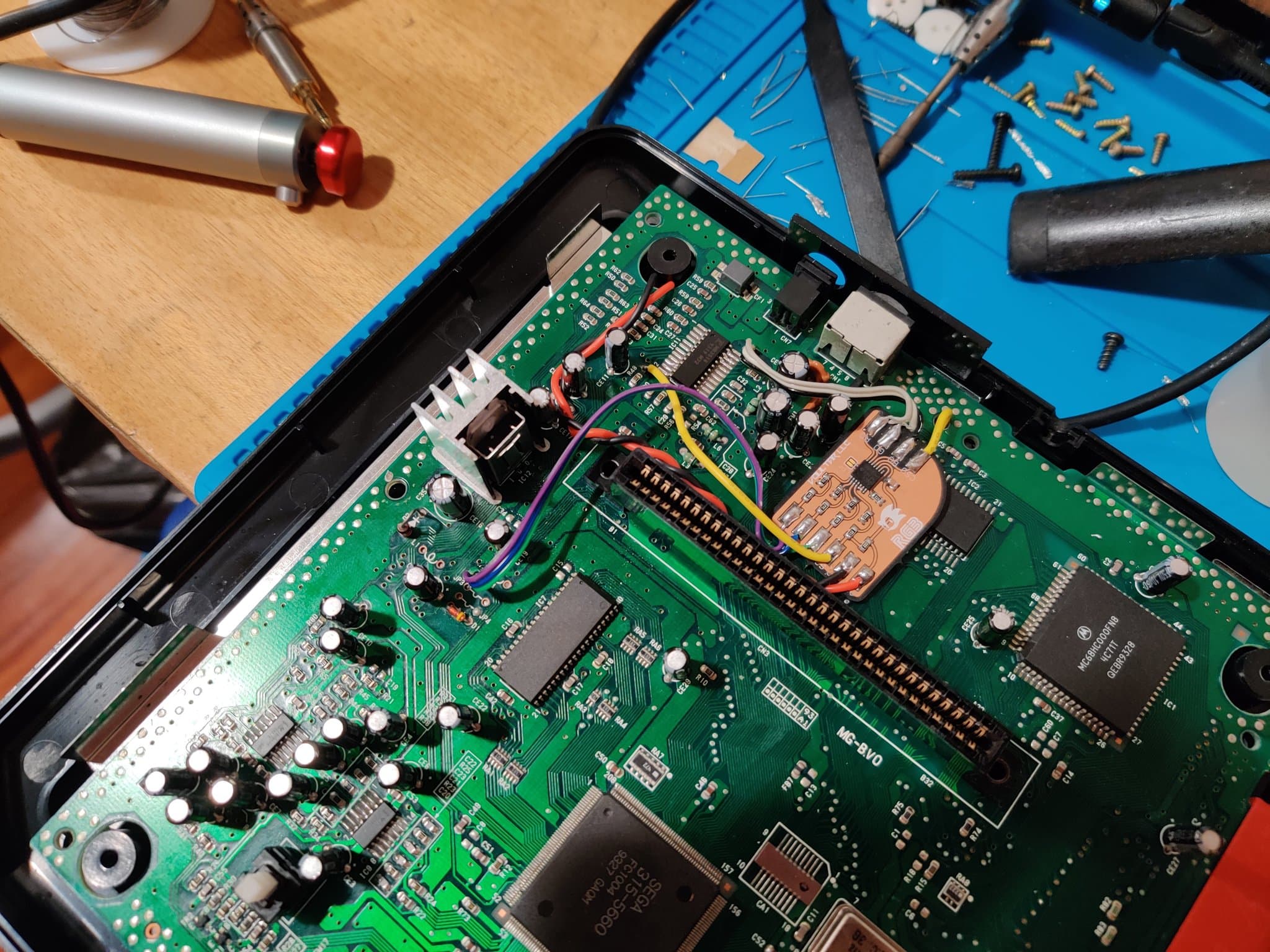 Soldered Mega Drive 2 RGB bypass mod installed on green PCB with colored wires, test setup on workbench with tools and components visible.