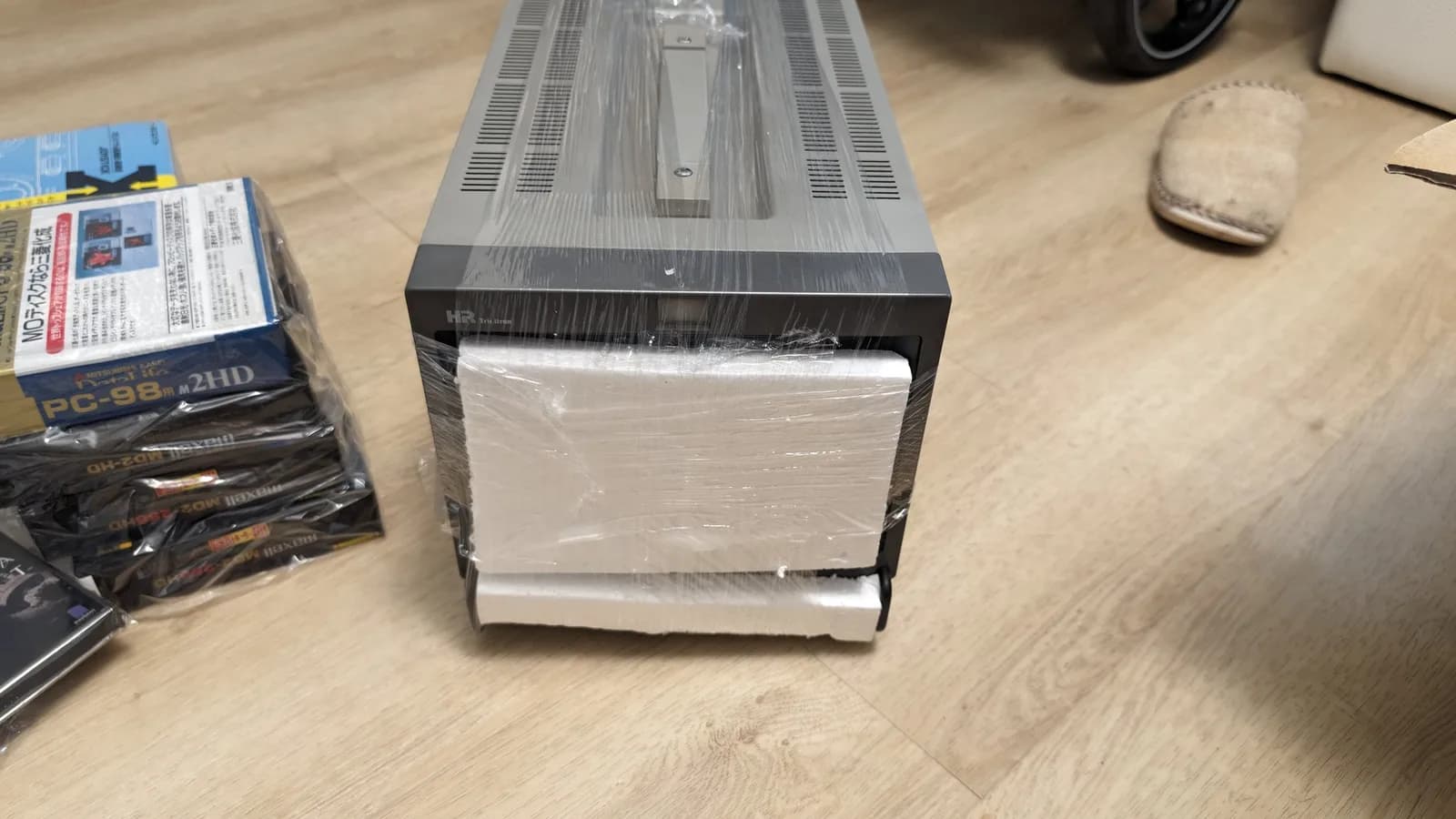 The Sony PVM-9045QD as it arrived from Japan, still in its shipping plastic wrap with styrofoam blocks over the screen, on a wood floor beside the floppy-disk boxes from the same haul.