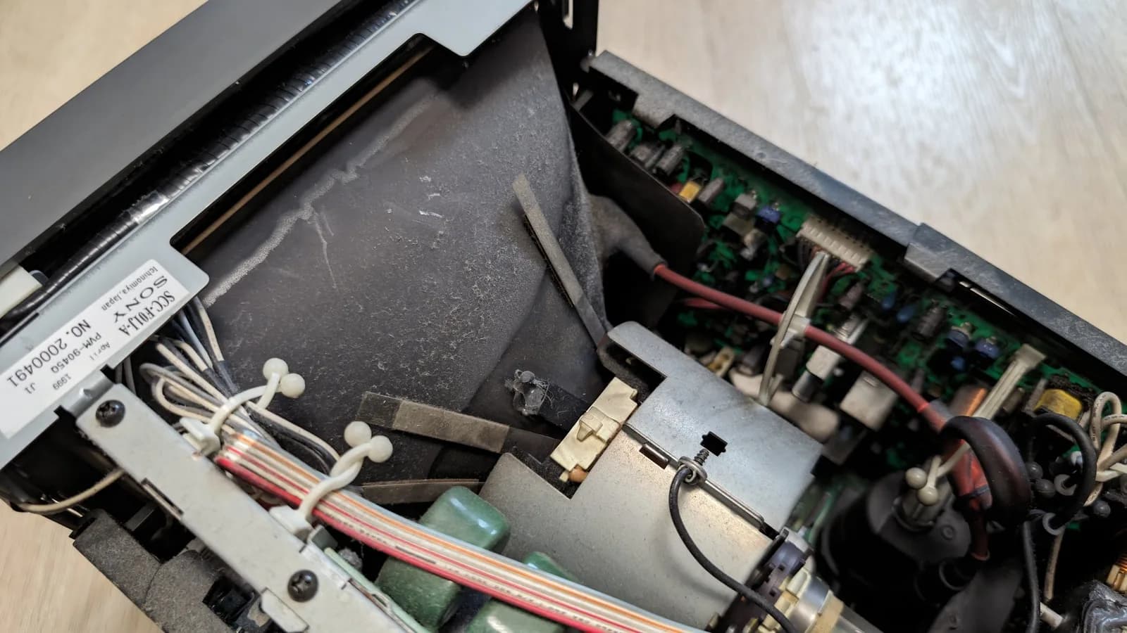 Inside the opened Sony PVM-9045QD (its factory sticker showing the serial number, model name and date visible), heavy dust over the CRT funnel and neck, cable-tied wiring, green capacitors and a circuit board alongside, before cleaning.
