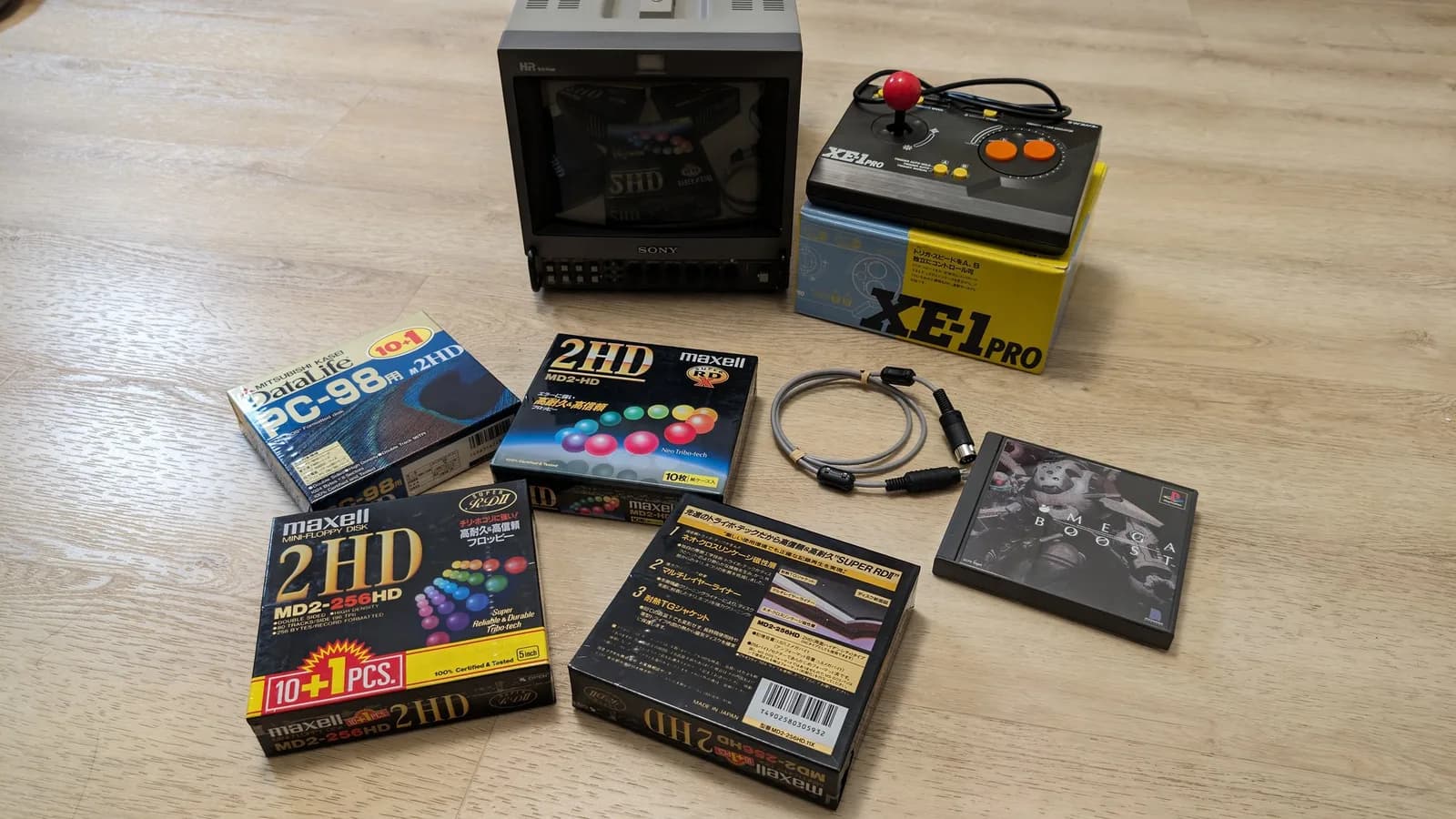 Sony PVM-9045QD (front view, screen off and reflecting the items in front of it) with a Japanese import haul on a wood floor: Maxell and Mitsubishi 2HD floppy-disk boxes, a Micomsoft XE-1 Pro joystick (for MSX/X68000) and its box, a MIDI cable, and the PS1 game Omega Boost.