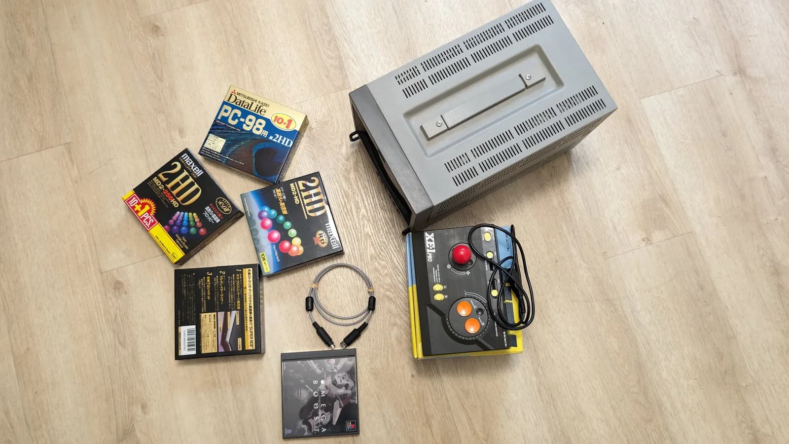 Overhead view of the Japanese import haul on a wood floor: the Sony PVM (top and carry handle visible), a Micomsoft XE-1 Pro joystick (for MSX/X68000) on its box, Maxell and Mitsubishi 2HD floppy-disk boxes, a coiled MIDI cable, and the PS1 game Omega Boost.