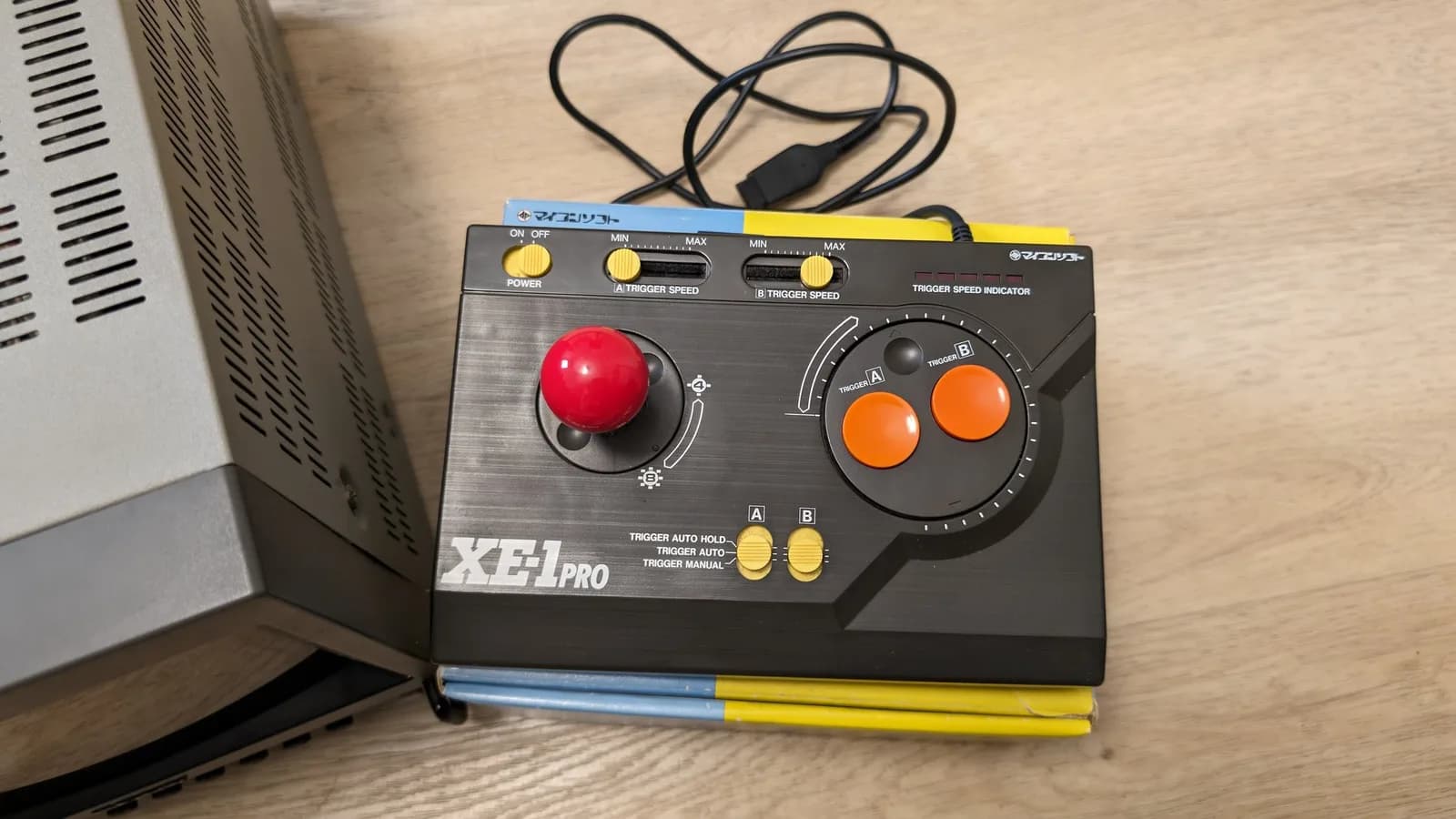Close-up of the Micomsoft XE-1 Pro joystick, black top plate with a red ball-top stick, two orange buttons and rows of trigger-speed switches, on its box next to the Sony PVM's vented side panel.