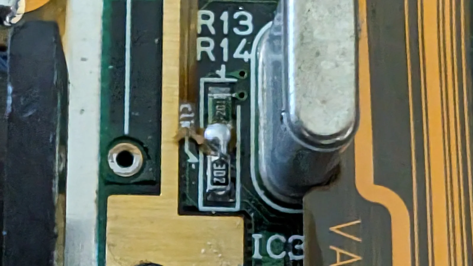 Macro close-up of the Game Gear mainboard at the clock signal point, between resistors R13 and R14 with IC3 alongside, showing the broken flex ribbon connector that cut the clock signal.