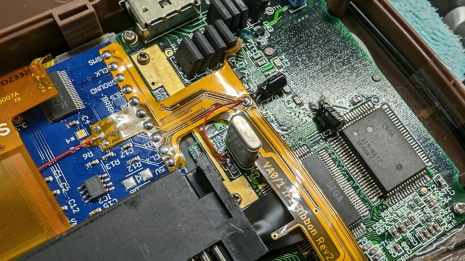 Close-up of the Game Gear mainboard repair: a red clock-line bodge wire runs from the clock point on the board, between two resistors, to the clock point on the flex ribbon, with UV curing solder mask at top right covering the TP10 pad. The blue IPS interface board sits alongside.
