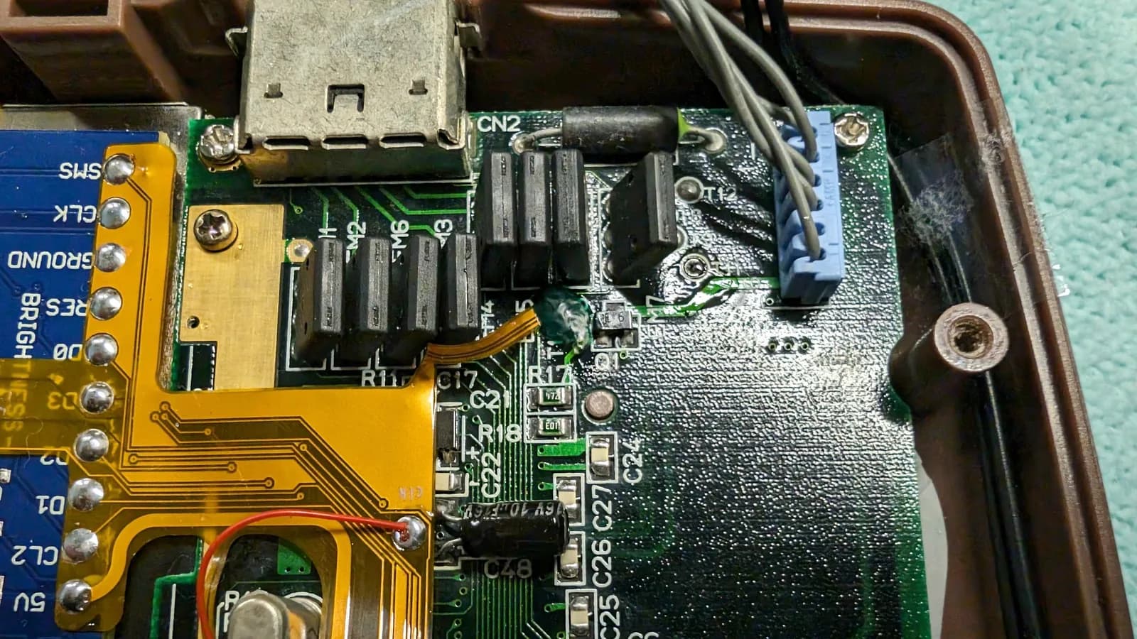 Front-facing close-up highlighting the clock-line repair on the Game Gear mainboard: UV curing solder mask over the TP10 pad, where the flex ribbon's clock line is soldered, the mask added to strengthen the joint and stop the ribbon tearing again. The red bodge wire, connector CN2, the orange flex and the blue IPS interface board are also in frame.