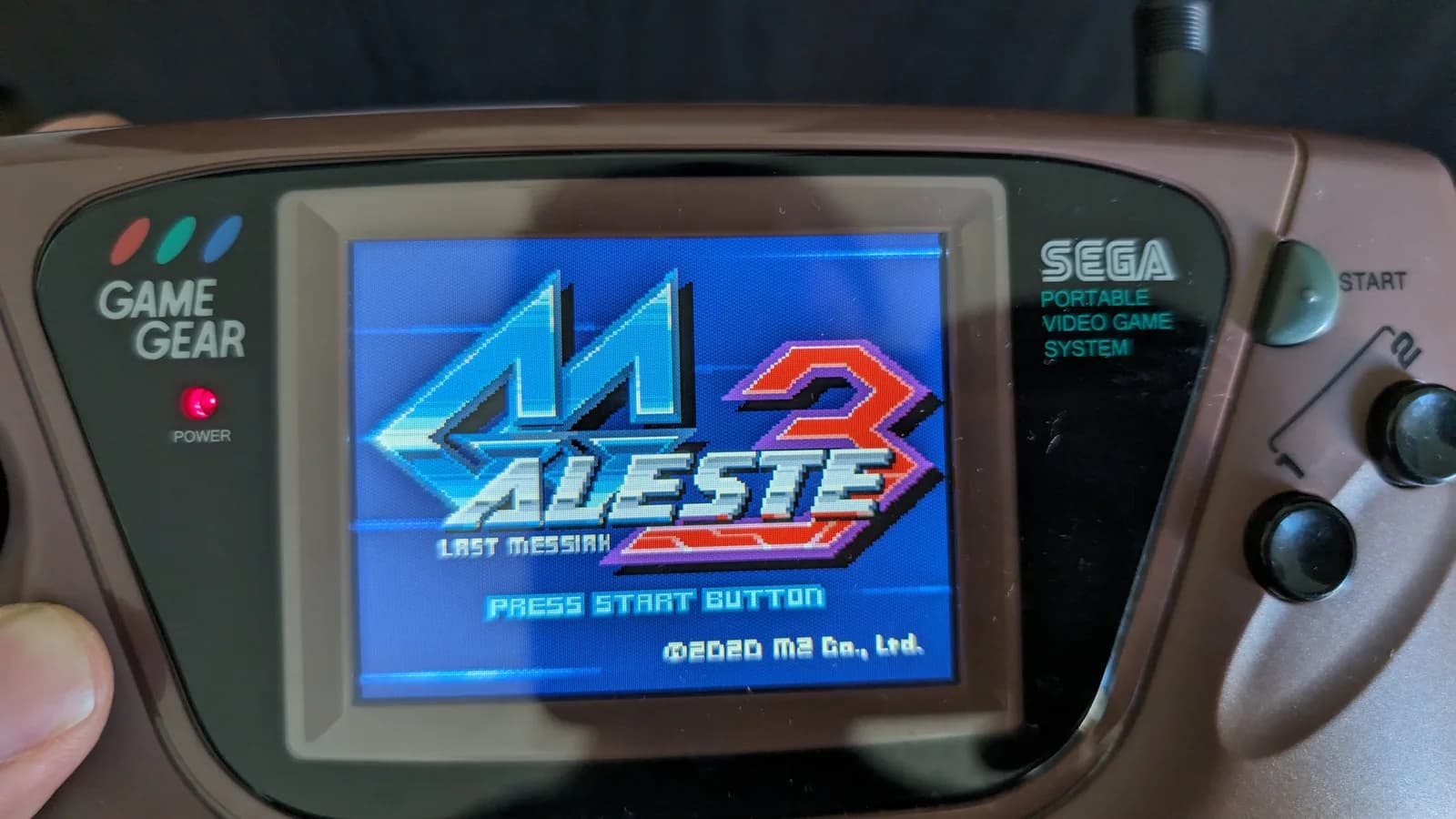 Brown Sega Game Gear held in hand, IPS screen restored and displaying the Aleste 3: Last Messiah title screen (PRESS START BUTTON, 2020 M2 Co., Ltd.) after the repair.