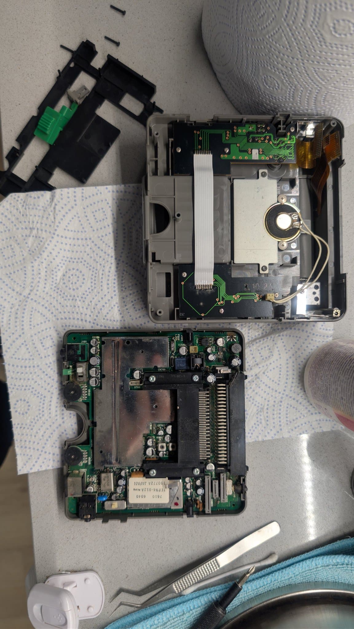 PC Engine LT split into its two case halves: the mainboard still seated in the bottom half, and the plastic frame, power button and GAME/TV slider lifted out to the top of the shot. Between them sits the top half, showing the speaker, the LCD's flex cables, and the two side control PCBs joined by solder-on ribbon wire.