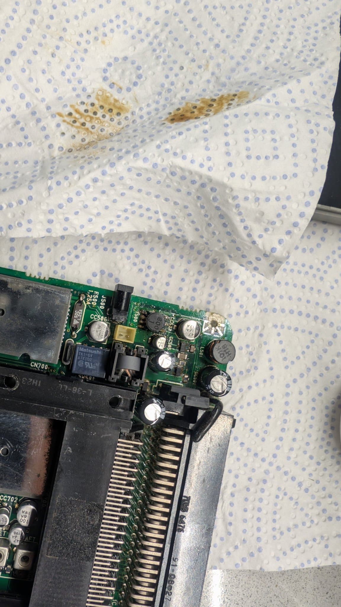 Close-up of the PC Engine LT board's power-input area: the T500 transformer, the input fuse, a Matsushita relay and the 5V linear regulator (24M05), with leaked electrolyte spread across the transformer, capacitors and SMD parts, and more pooled on the paper towel above.