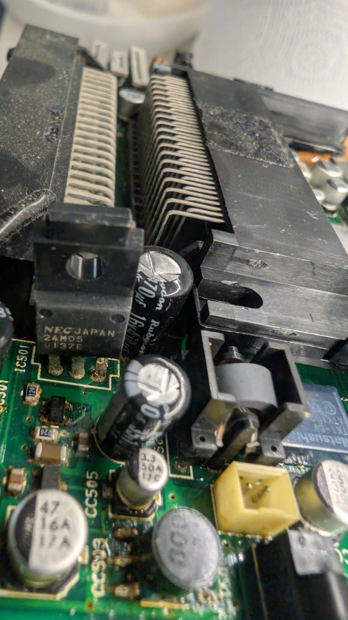 Close-up of the PC Engine LT board: a TO-220 regulator marked NEC JAPAN 24M05 beside a Rubycon 470uF 16V capacitor, with leaked electrolyte across the regulator, the surrounding capacitors and SMD components, and the T500 transformer at the bottom of the shot.