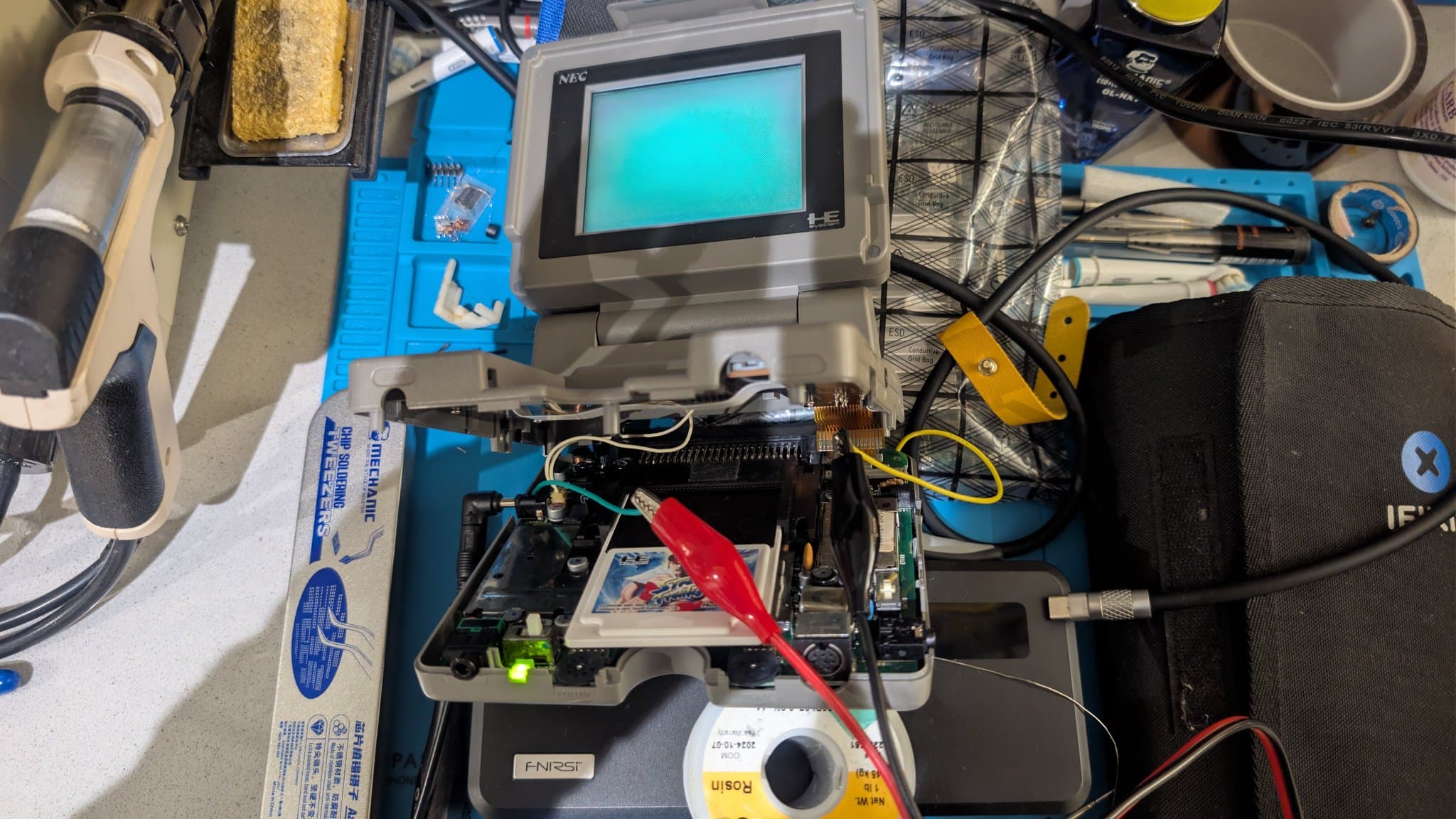 Opened NEC PC Engine LT on a blue mat, powered from the bench PSU with red and black alligator clips probing the exposed lower PCB and HuCard slot, amid repair tools. The LCD is powered on, its image washed out at this off-axis viewing angle.