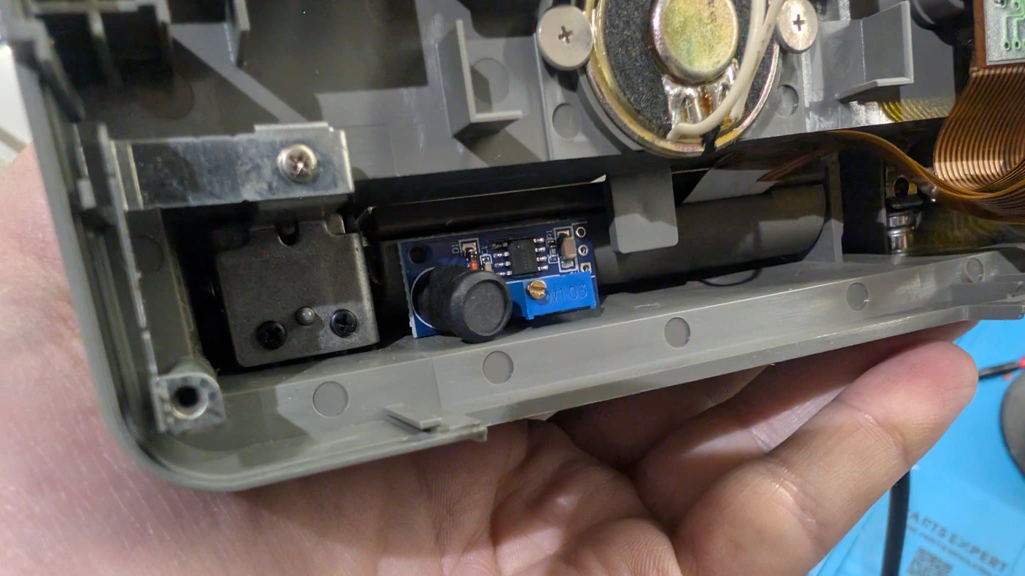 Hand holding the PC Engine LT top shell with a small blue boost and inverter module (trimmer pot and capacitor) fitted into the shell recess, near the speaker and coil wiring.
