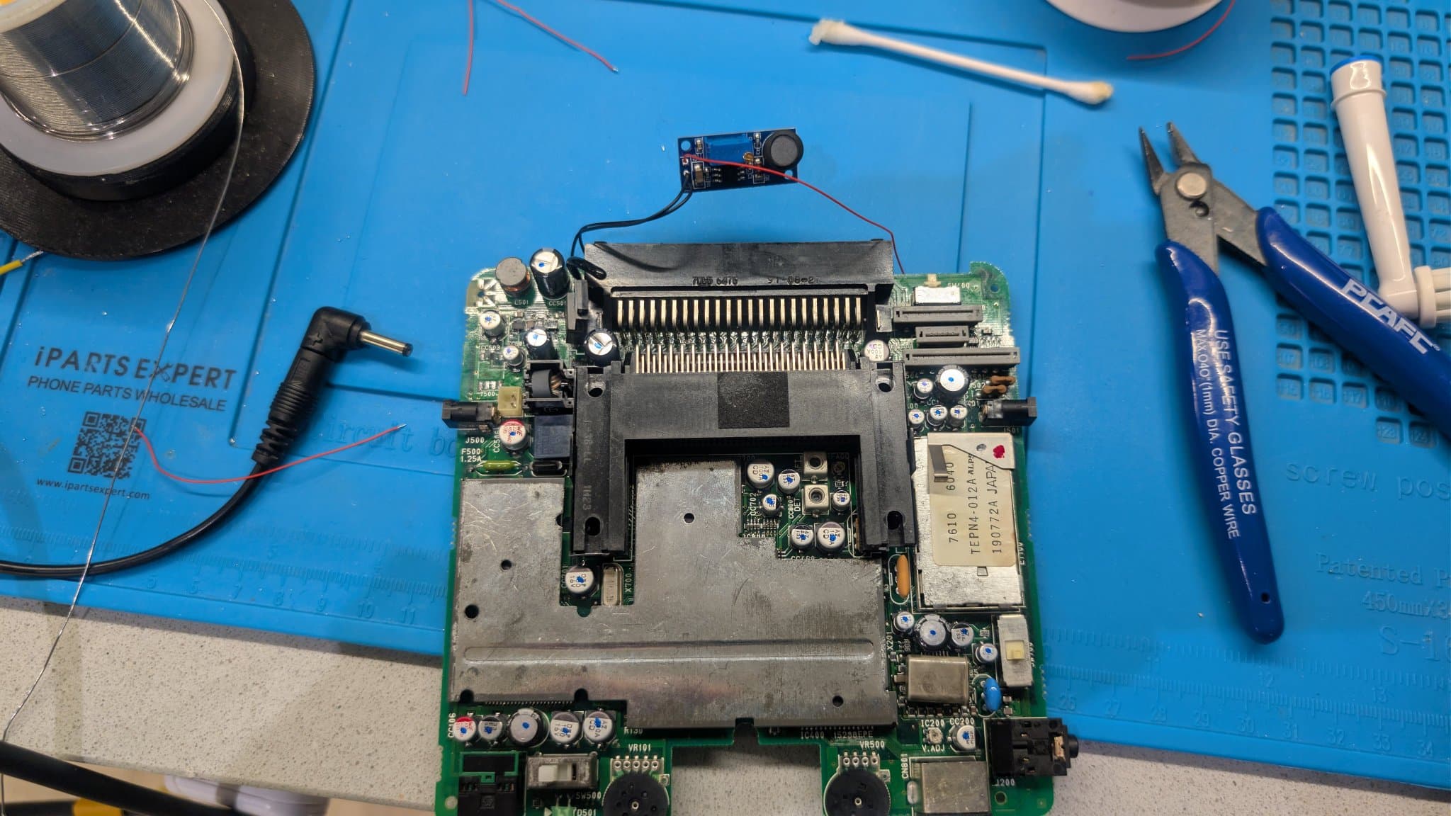 PC Engine LT motherboard on a blue mat with the small blue boost and inverter module wired in at the top edge to stand in for the failed T500 transformer, beside the HuCard slot and repair tools.