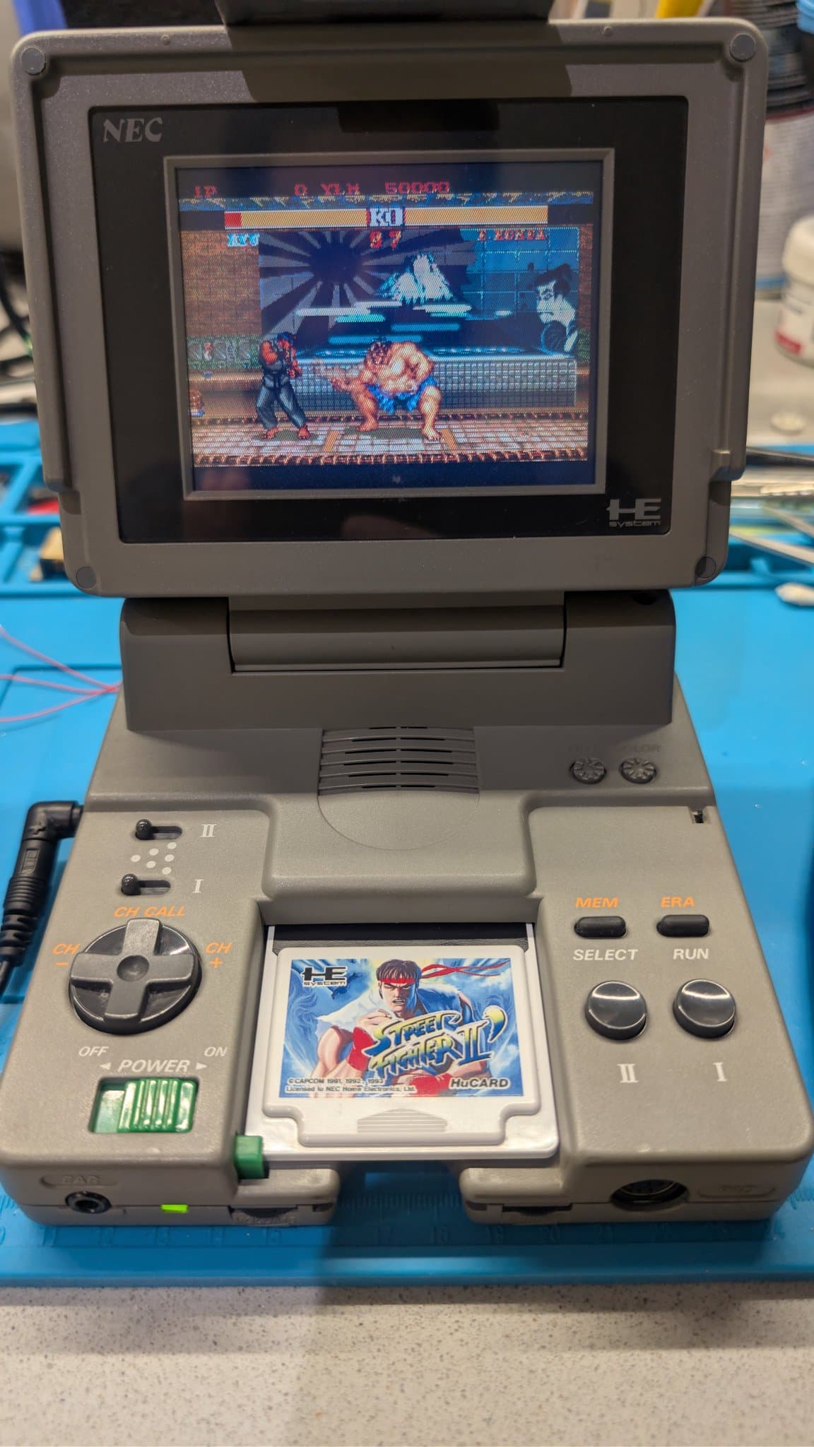 Assembled NEC PC Engine LT with the lid up showing Street Fighter II gameplay, Ryu fighting E. Honda, and a matching Street Fighter II HuCard in the front slot, on a blue mat.