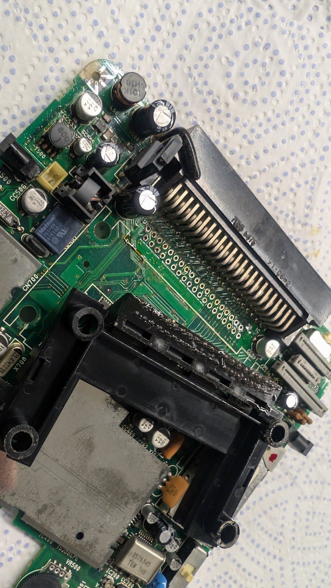 Component side of the PC Engine LT mainboard at the 5V regulator and the power input jack, with capacitors and electrolyte grime spread everywhere. The HuCard slot connector has been desoldered and lies in view, both its underside and its former footprint on the board caked with electrolyte and dirt.