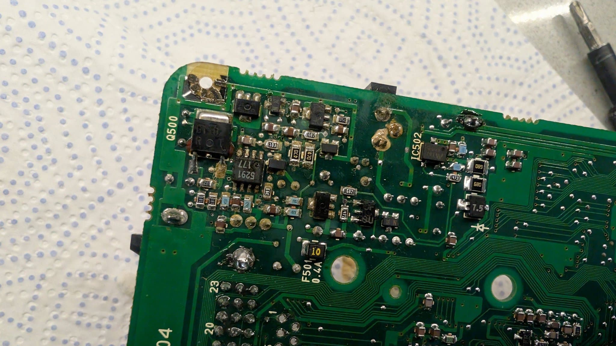 Close-up of the PC Engine LT board's SMD power section: the 8-pin 6.5V regulator marked 5291 (M5291FP) sitting upside down in the shot, next to the Q500 position, with electrolyte contamination on the solder of nearly every component in the area and a screwdriver tip entering from the corner.