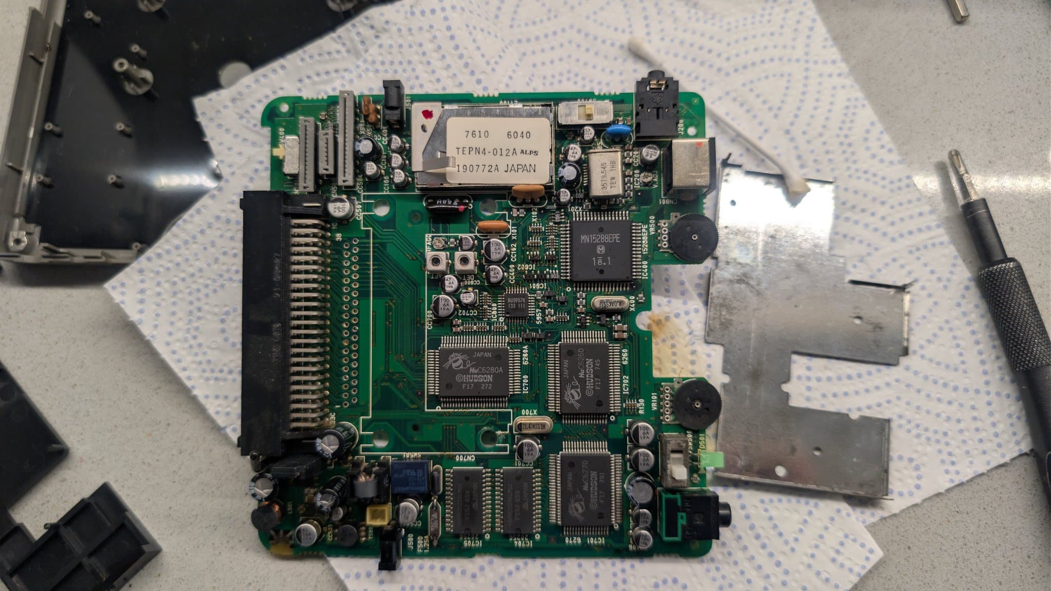 Top-down view of the cleaned PC Engine LT mainboard with its ALPS metal-can module, the Hudson HuC6280A and other ICs, the HuCard slot and the power jacks at the board edges, beside the shell and tools.