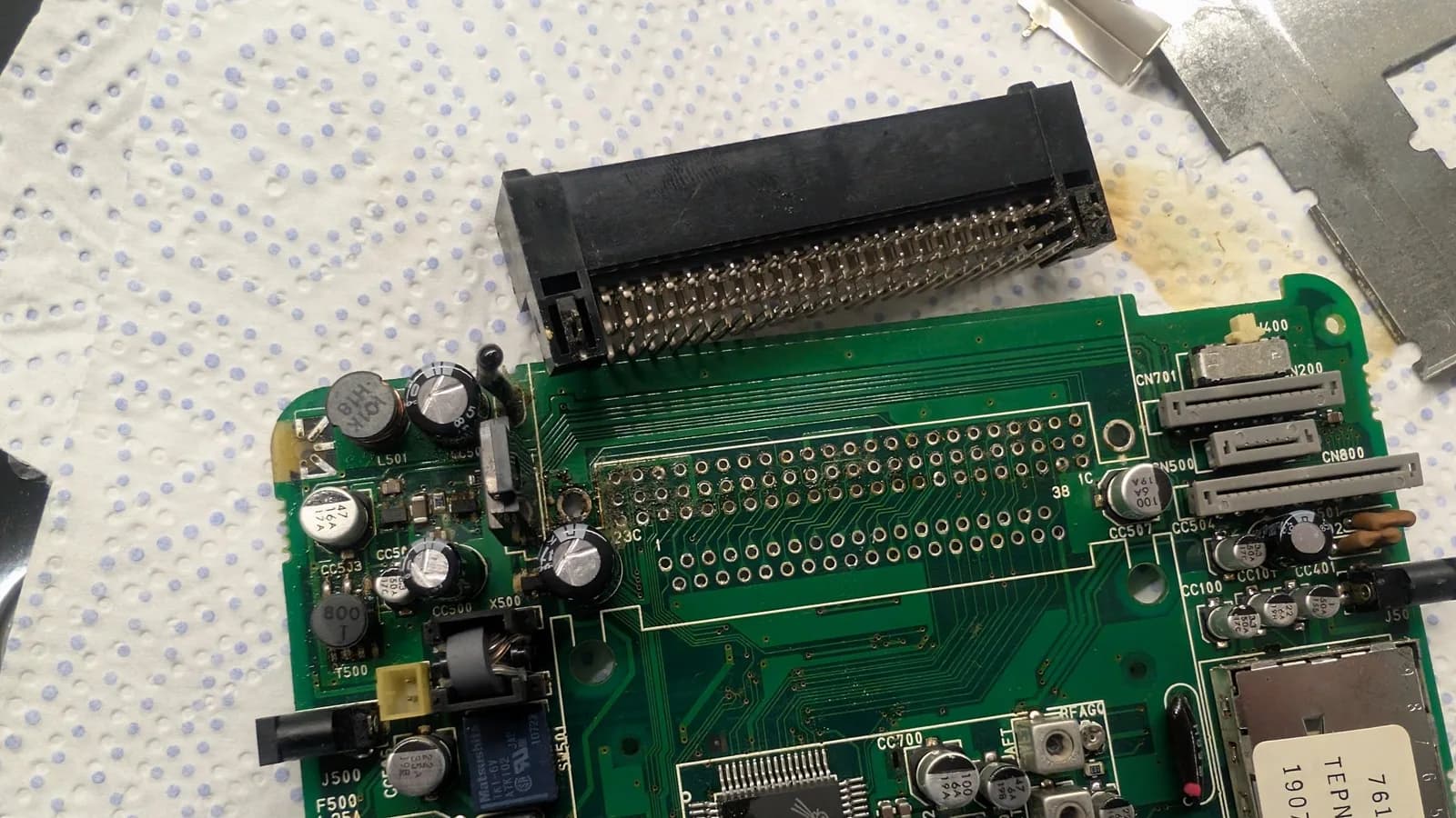 PC Engine LT board (component side) on a paper towel with the Ext Bus connector desoldered and resting above it, exposing the electrolyte and grime caked on the board where the connector had sat.