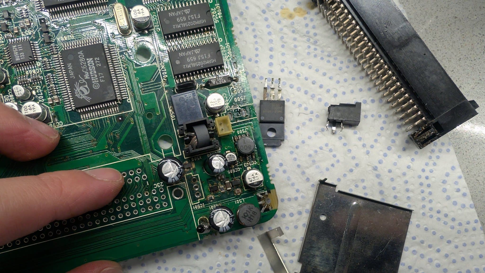 PC Engine LT board held in hand with the Ext Bus connector, the 5V regulator (24M05) and the power input jack removed and laid on a paper towel nearby.