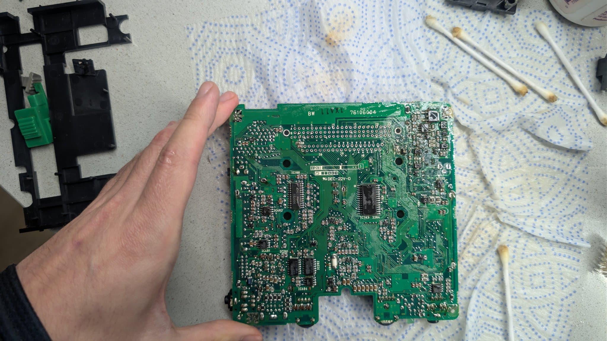 Hand holding the NEC PC Engine LT mainboard by its edge, the board flipped to show its solder side after cleaning with white vinegar, with cotton swabs and the removed plastic frame (carrying the power slider and GAME/TV slider) behind it.