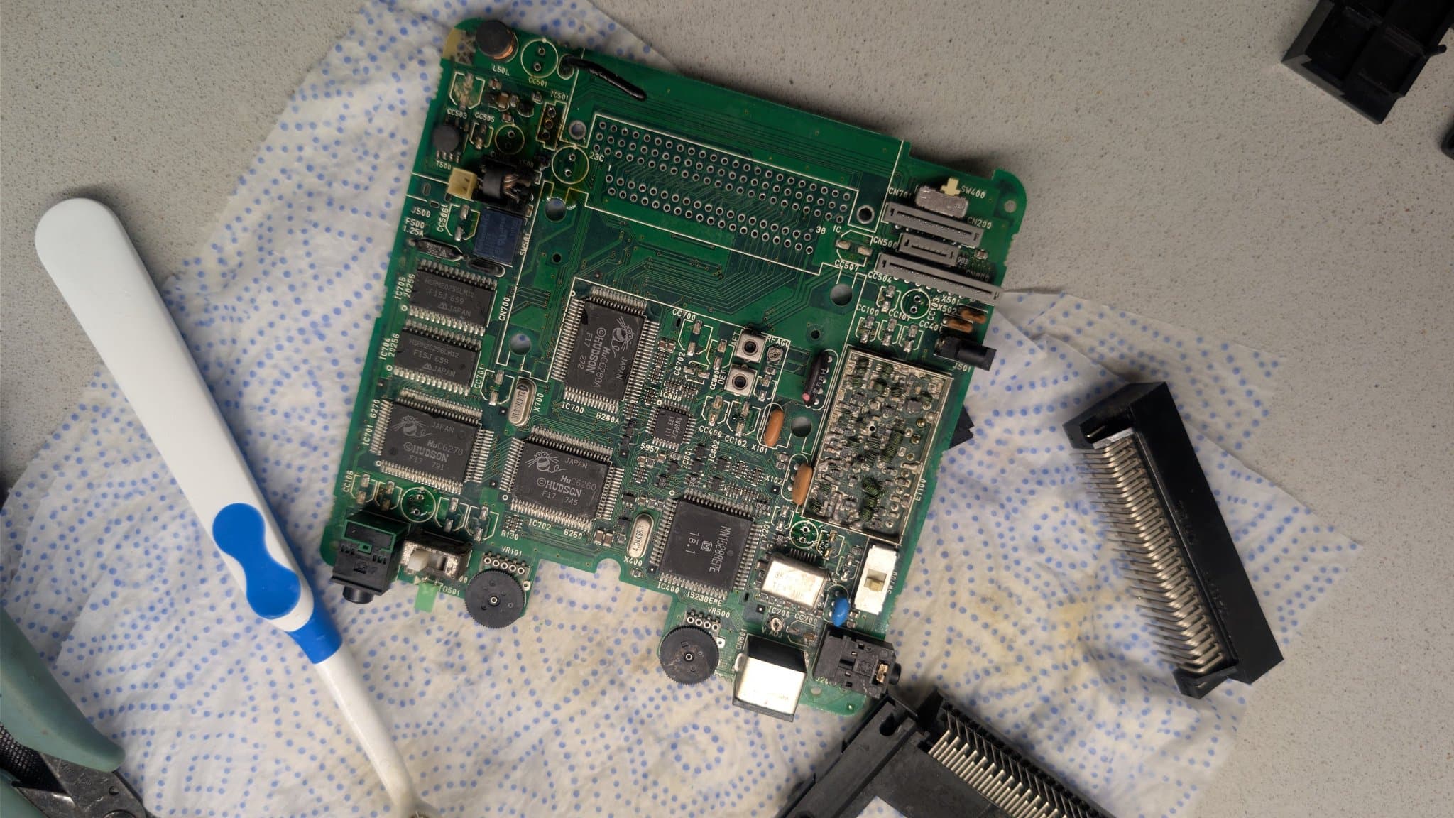 PC Engine LT board (component side, cleaned with white vinegar and with its capacitors already removed) on a paper towel, a cleaning brush at left and the removed HuCard slot and Ext Bus connector at right.