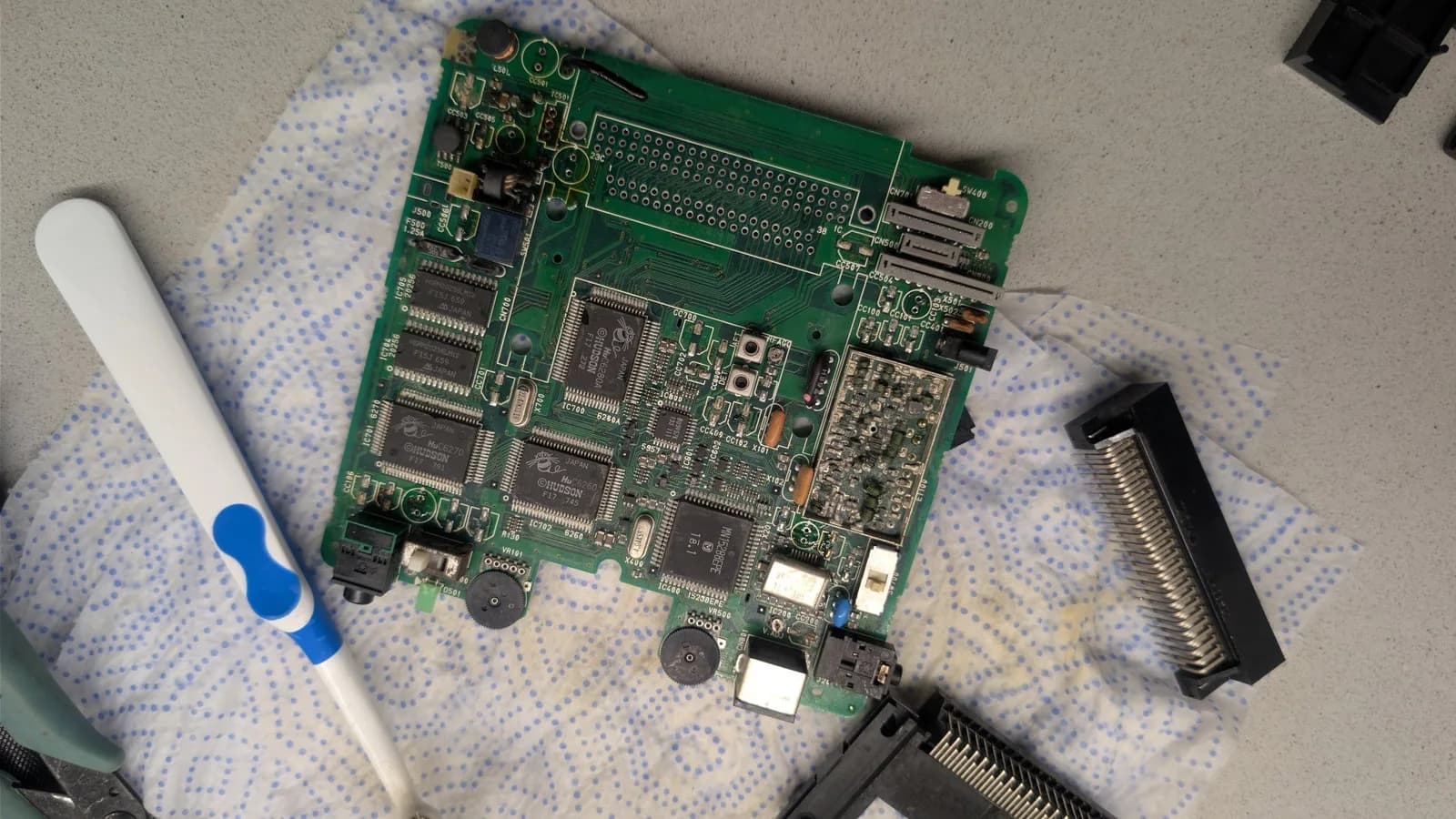 PC Engine LT board (component side, cleaned with white vinegar and with its capacitors already removed) on a paper towel, a cleaning brush at left and the removed HuCard slot and Ext Bus connector at right.