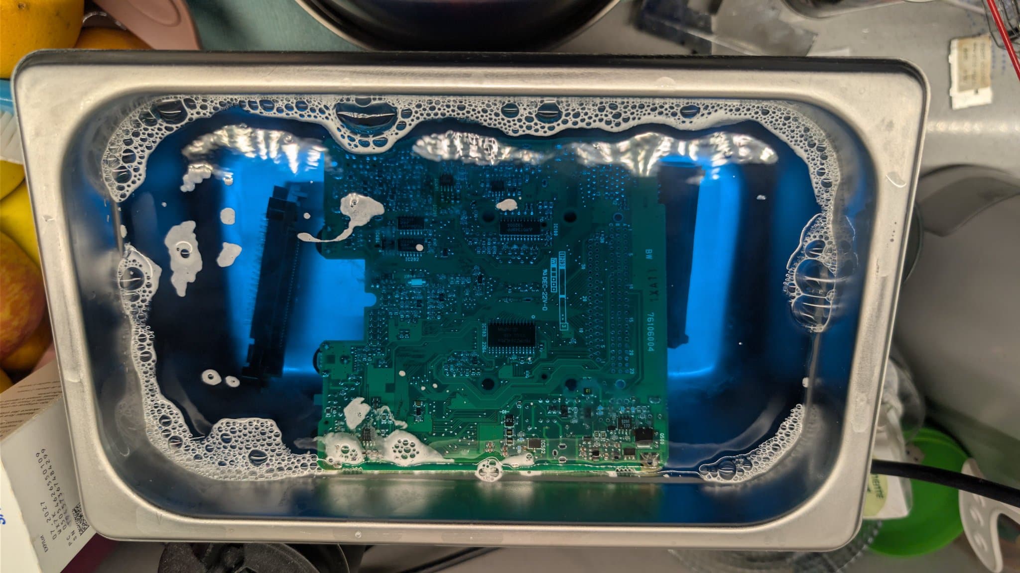 NEC PC Engine LT board (solder side) submerged in a stainless steel ultrasonic cleaner tank with foamy water.