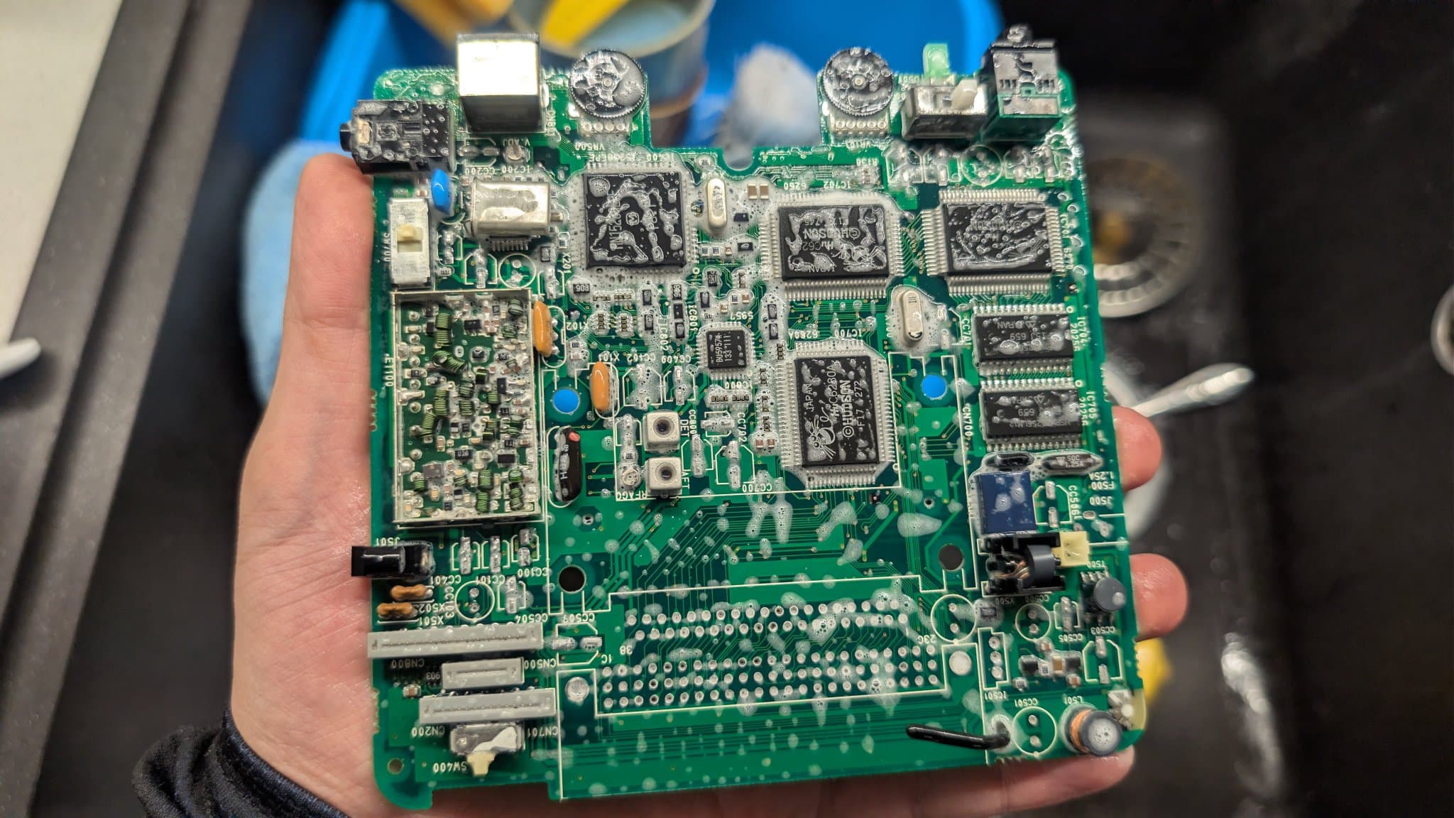NEC PC Engine LT mainboard (component side) held in hand, drenched and foamy after being scrubbed coming out of the ultrasonic bath, its chips and small components on the green PCB.