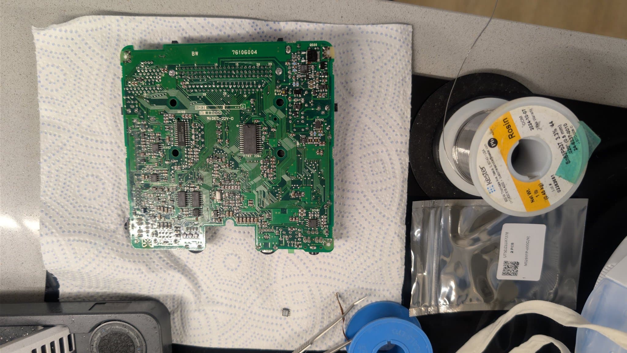 NEC PC Engine LT mainboard with a new 6.5V regulator (M5291FP) installed and the old one resting on the paper towel at the bottom of the shot, near tweezers and desoldering braid, plus a Kester solder spool and small bags of replacement parts (one holding spare M5291FP units like the one just swapped).