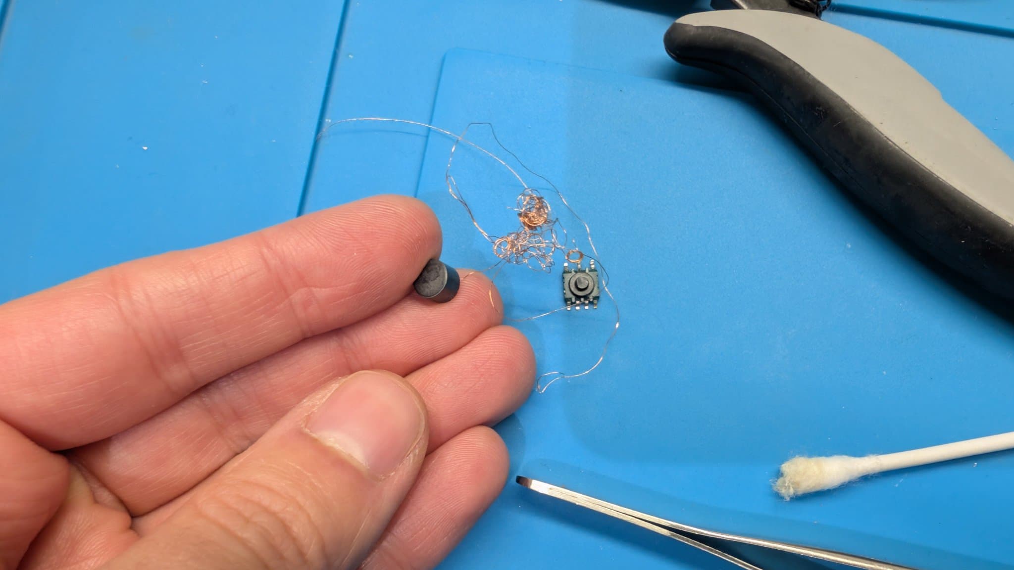 Hand holding the destroyed T500 transformer beside its unwound copper winding and a small core, with pliers and tweezers nearby during the PC Engine LT repair.