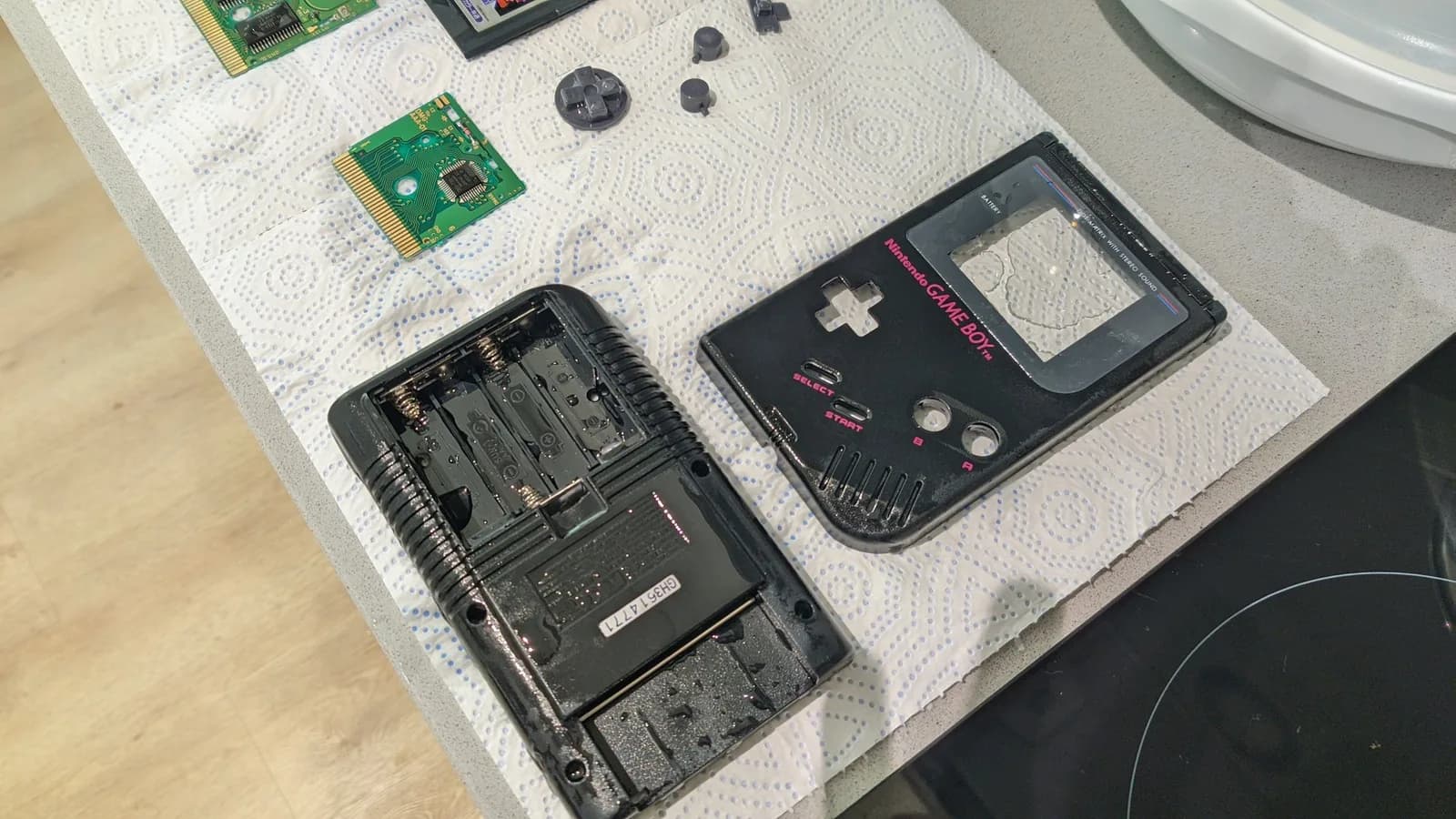 Black Play It Loud Game Boy DMG-01 disassembled on a white, blue-dotted paper towel: the two shell halves (front fascia with the Nintendo GAME BOY logo and the lens film still on, and the rear shell with its empty battery compartment) and the loose grey D-pad and A/B buttons freshly cleaned and still wet, with two green Game Boy cartridge boards (the Tetris carts being refurbished) alongside.