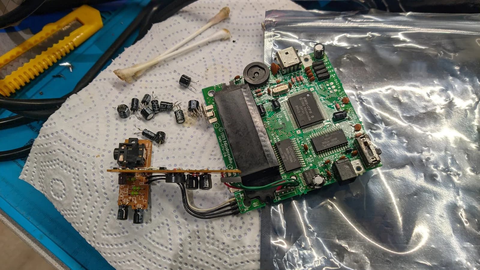 Game Boy DMG-01 main board (DMG-CPU under its metal shield, link port and speaker) on an antistatic bag, the old electrolytic capacitors and transistors removed onto the paper towel beside it. The DMG's two stock sub-boards are still attached to it by their factory wiring: the audio board with its 3.5mm headphone jack (three black and one white lead) resting on the towel, and the power board seen edge-on as a thin sliver of capacitors and components, propped up on its red and three green wires.