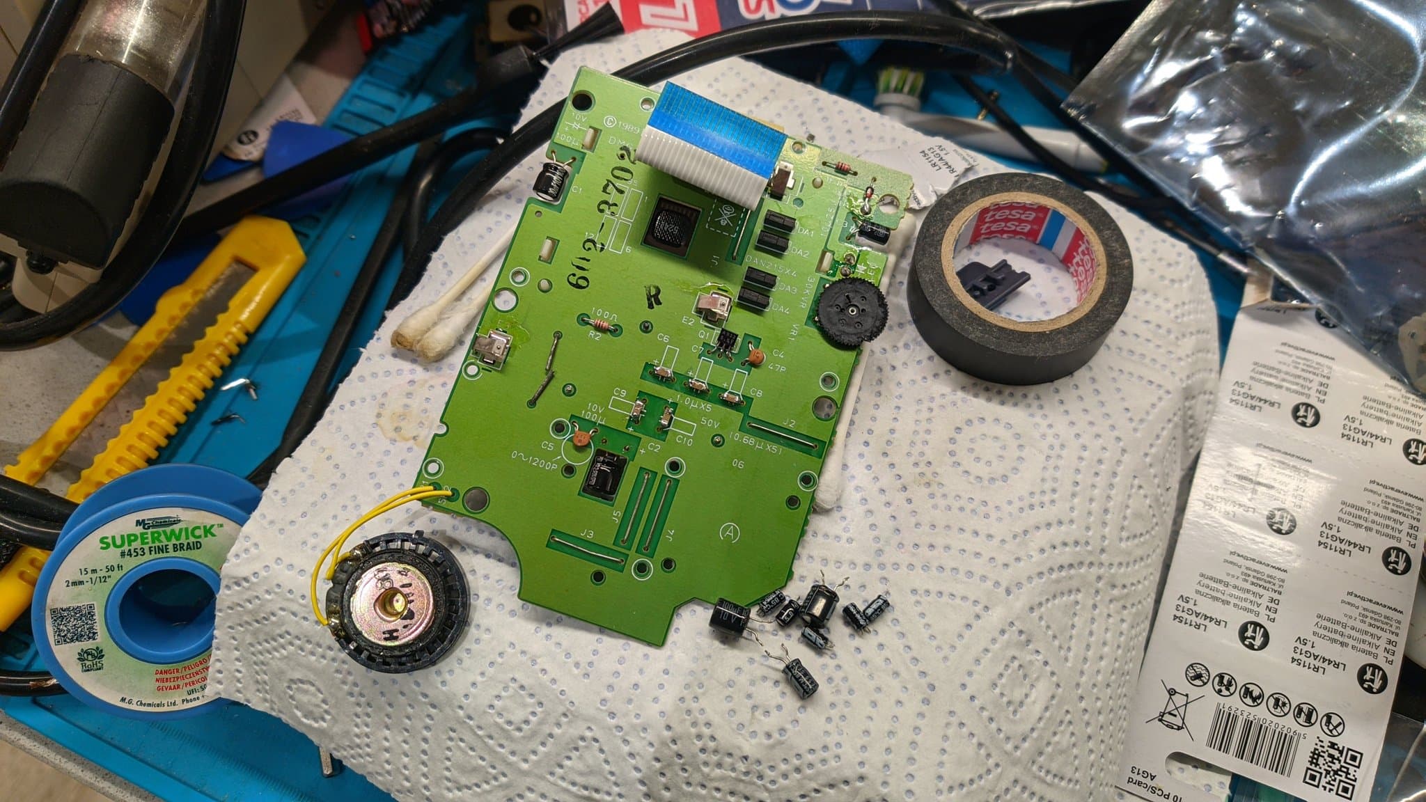 Green Game Boy motherboard with new capacitors installed, surrounded by soldering tools, desoldering wick, and documentation during restoration work.