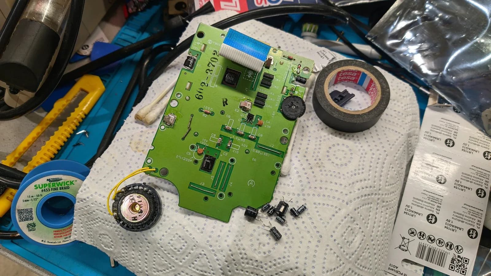 Green Game Boy DMG-01 LCD and regulator board, recapped from a Console5 capacitor kit with the old electrolytics removed beside it; at C6 to C10 the kit's replacements are ceramic caps soldered onto the remains of the old ones (a Console5 ease-of-assembly change). A roll of Superwick desoldering braid, hand tools, electrical tape and LR44 button-cell battery packaging are alongside.