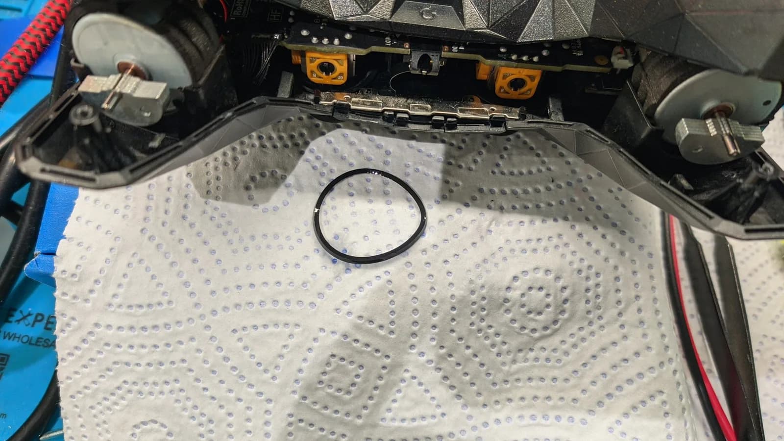 The Xbox 360 optical drive tray's belt on a white paper towel after boiling, its circular shape almost fully restored, an Nvidia Shield controller above it and the shell partially opened.