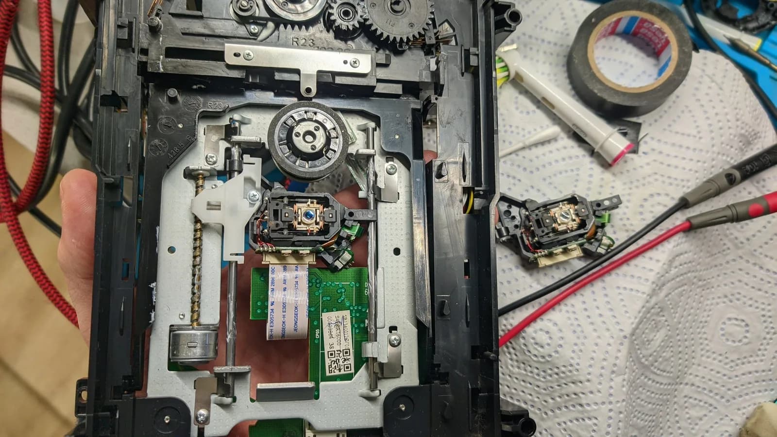 The Xbox 360 disc drive with the new laser installed and the drive cleaned and lubricated, the old laser off to the side next to multimeter probes.