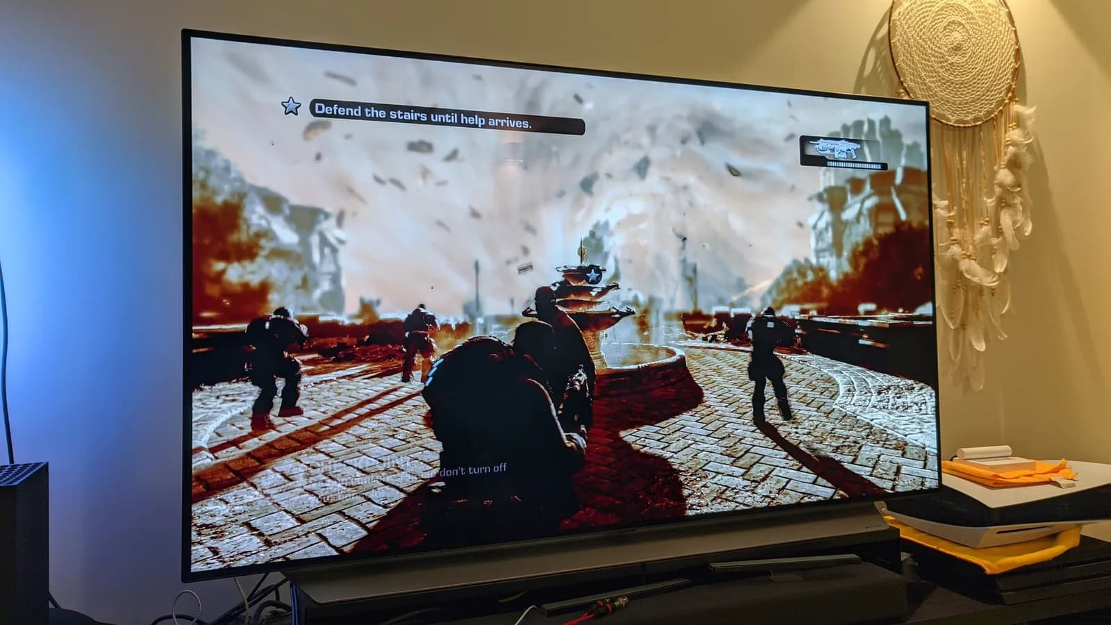 The Xbox 360 running Gears of War after the repair, the on-screen HUD reading the objective Defend the stairs until help arrives.