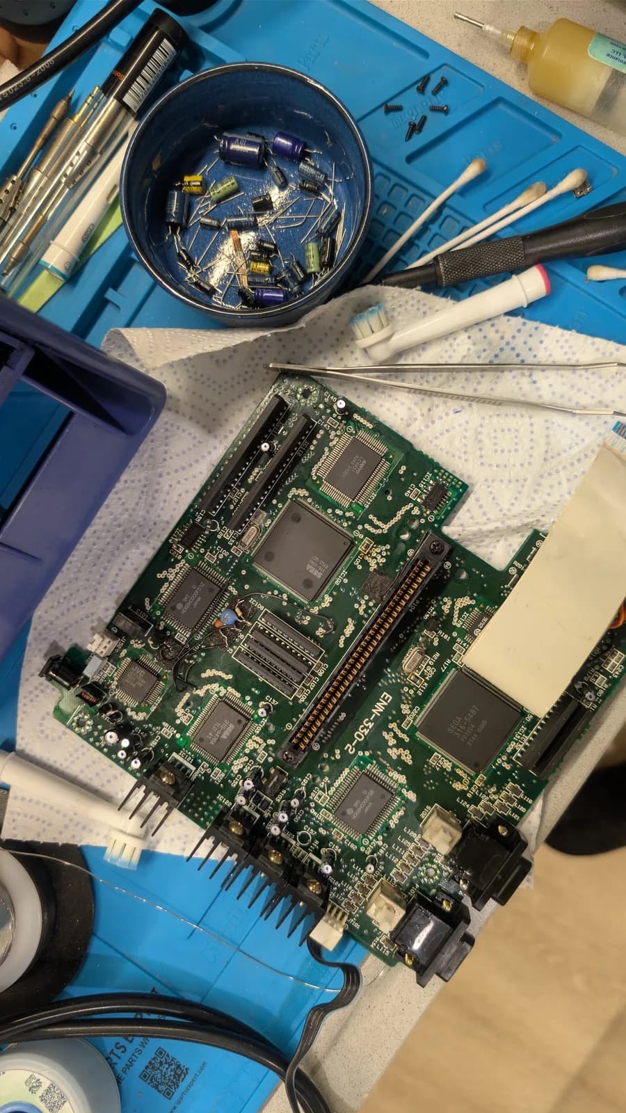 Sega Wondermega Victor RG-M1 board recap in progress: green circuit board on antistatic mat surrounded by capacitors in bowl, soldering iron, desoldering tools, and brushes on blue work mat.