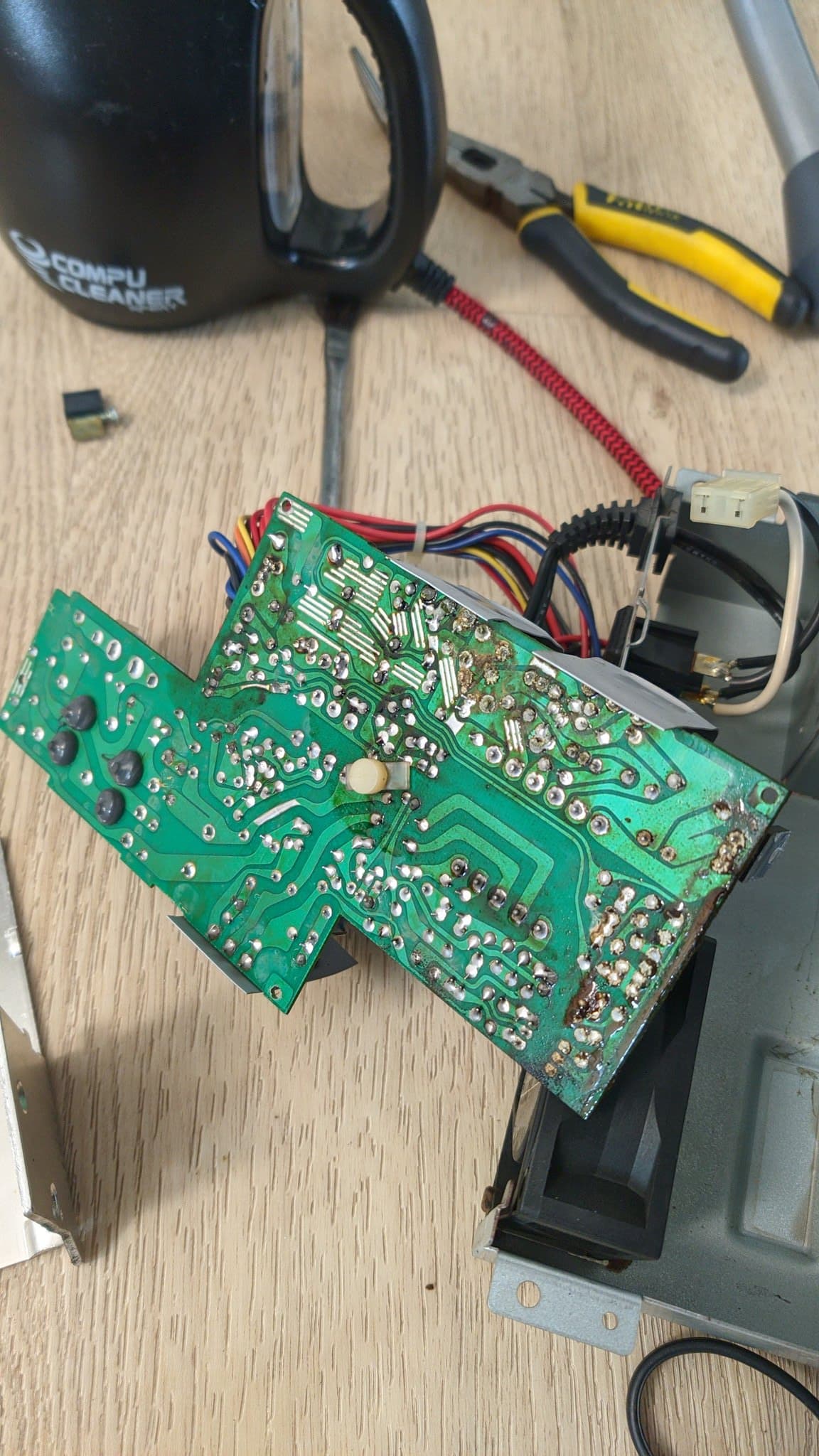 The underside of the Sharp X68000 Pro II power supply PCB, rested on top of the PSU's metal housing on the floor and being cleaned with a brush and a CompuCleaner air blower; massive electrolyte contamination covers the board, with darkened regions of the PCB and darkened solder joints, grime pooled where the electrolyte ran, and one edge of the board very dark from the contamination.