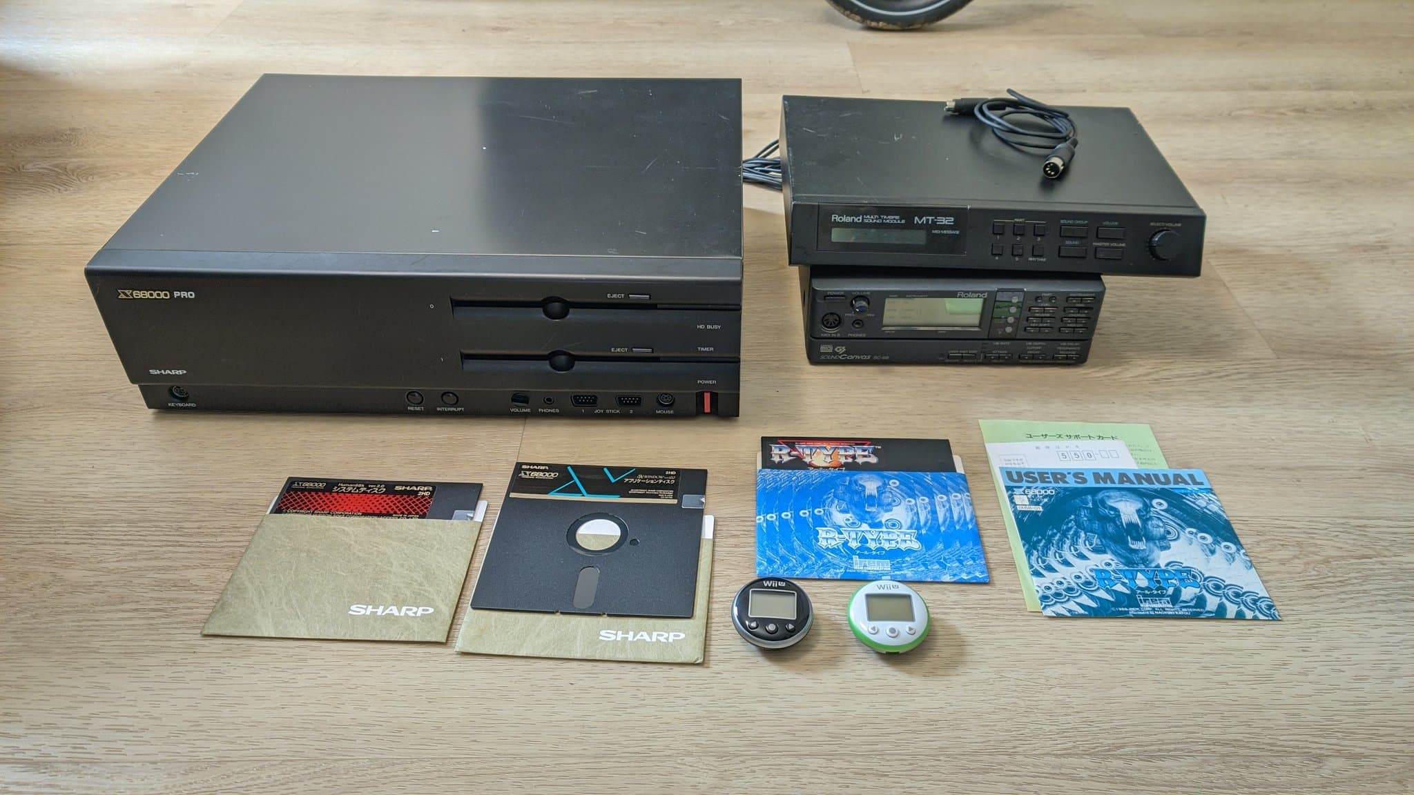 The haul newly arrived from Japan on a wooden surface: the broken Sharp X68000 Pro II, the Roland MT-32 and SC-88 MIDI sound modules, two Wii U pedometers, floppy disks, software and manuals.