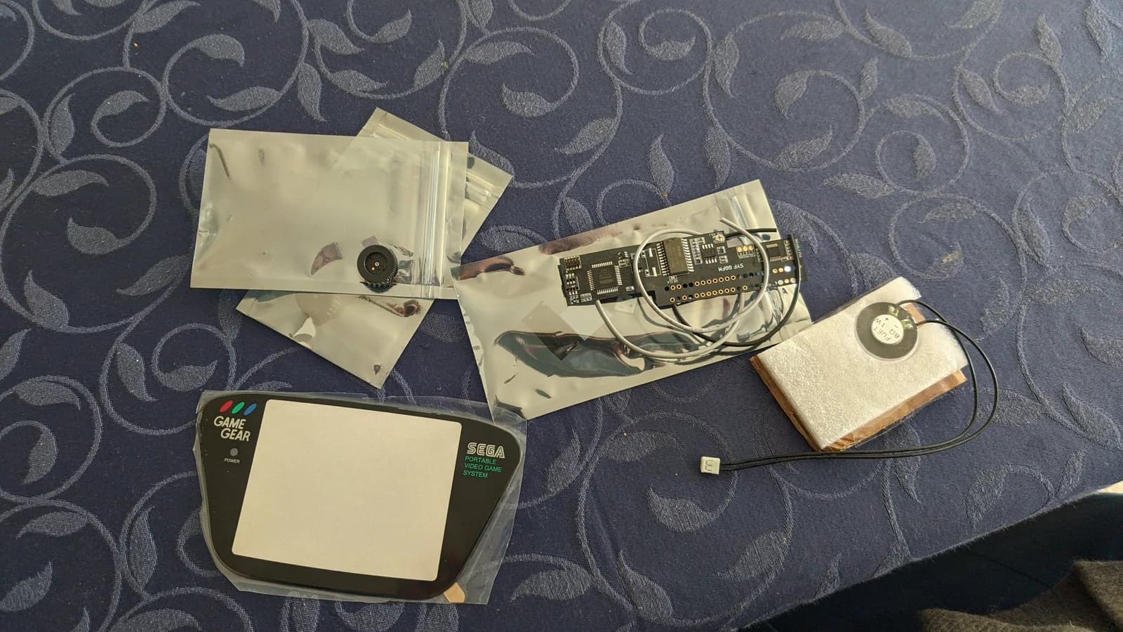 Game Gear mod parts laid out on a patterned surface: the round glass lens and other parts in antistatic bags, the FM sound module PCB with its wiring, a replacement speaker, a volume trimmer pot, and the front screen lens with the Game Gear and Sega markings.