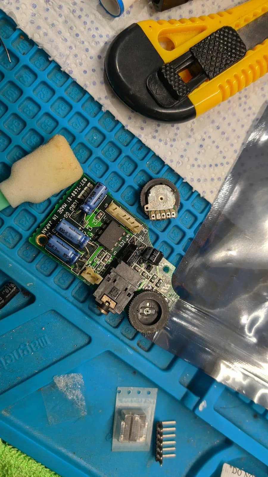 The Game Gear's sound board after a recap, with the new volume trimmer pot installed and the old trimmer pot removed and lying beside it, fresh electrolytic capacitors on the board, on a blue work mat with a box cutter and sponge nearby.