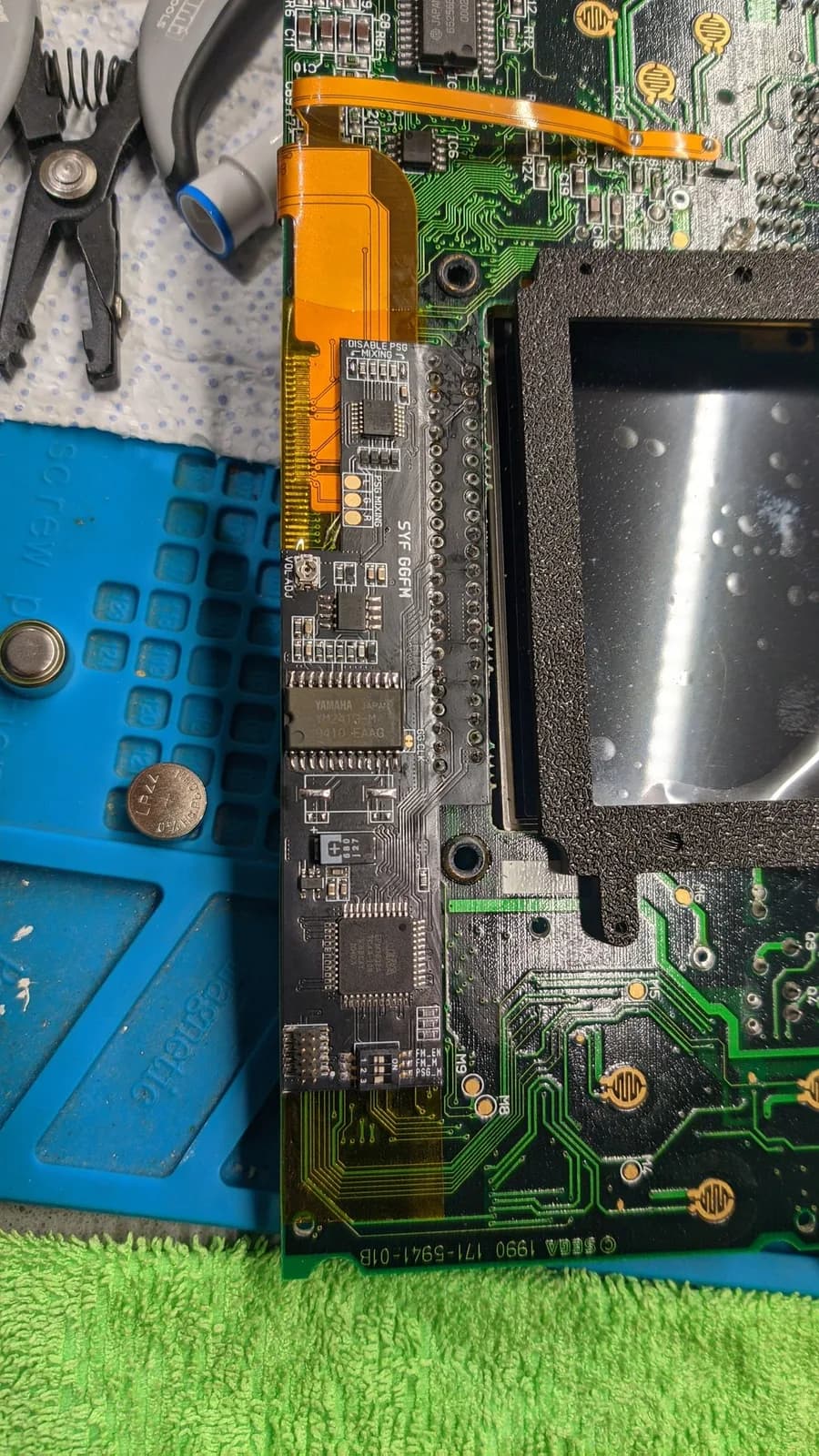 Close-up of the Game Gear interior with the FM sound module installed in place, the orange IPS flex cable routed past the black screen-frame opening nearby.
