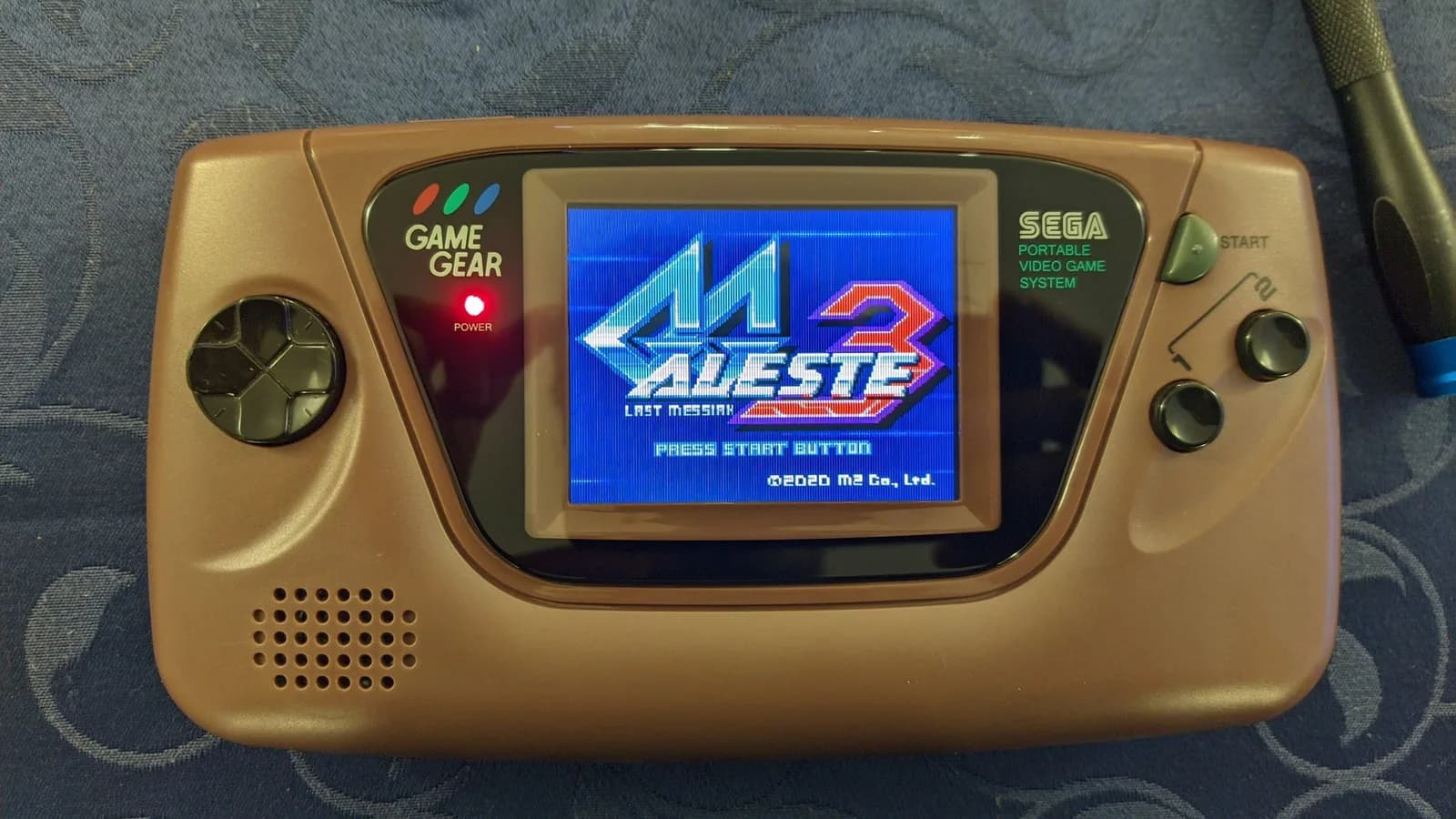 Brown Sega Game Gear with the IPS display mod showing the Aleste 3: Last Messiah title screen (PRESS START BUTTON, 2020 M2 Co., Ltd.); red power LED lit, speaker grille and d-pad visible.