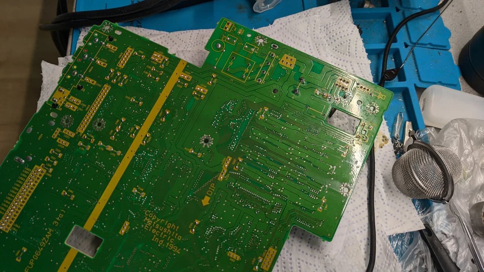 The solder side of the Panasonic FZ-1 mainboard during the recap ("Copyright Matsushita Electric Ind.1994", a "DIP" arrow), its through-hole solder joints exposed, with tweezers and a metal mesh tea-ball nearby.