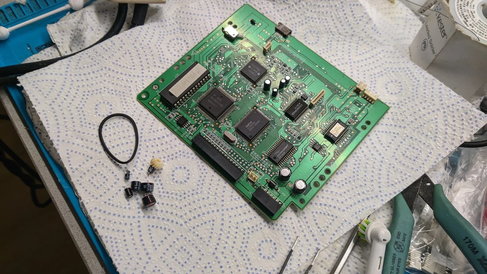 The 3DO FZ-1 disc-drive PCB during its recap, with the removed old capacitors, the replaced rubber drive belt (a black loop) and the broken yellowed plastic gear from the laser-sled shaft (replaced with a new 3D-printed one) in front, plus flush cutters, a brush and tweezers.