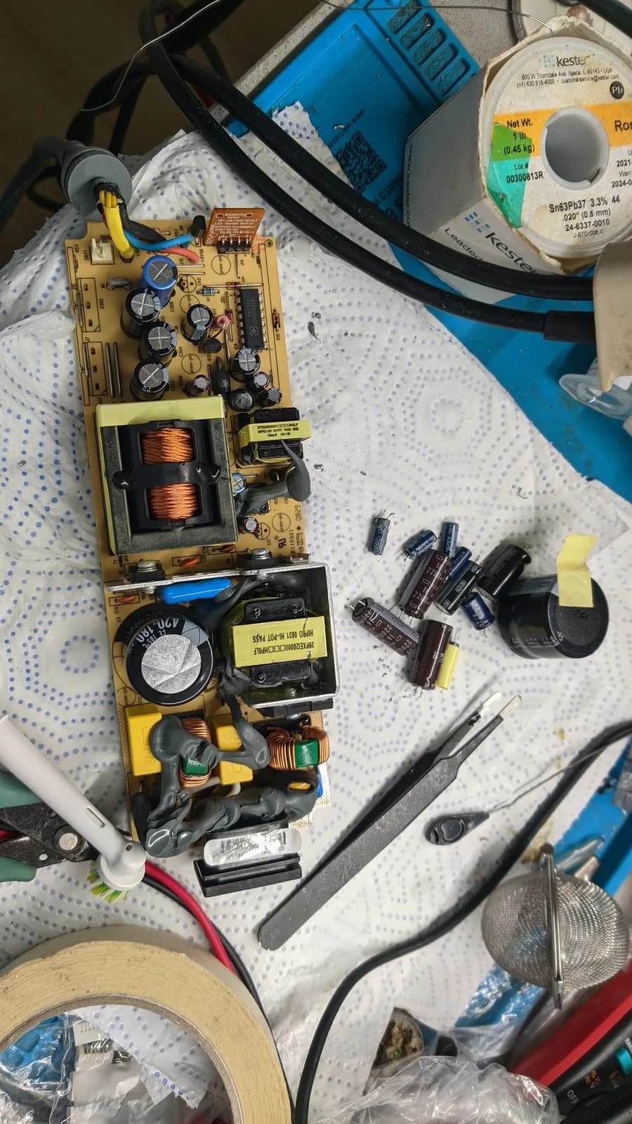 Xbox 360 S power supply board out of its case, component side up during the recap: electrolytic capacitors and wound transformers on the board, the old removed capacitors lying on the paper towel beside it, and a spool of solder, tweezers and cutters nearby.