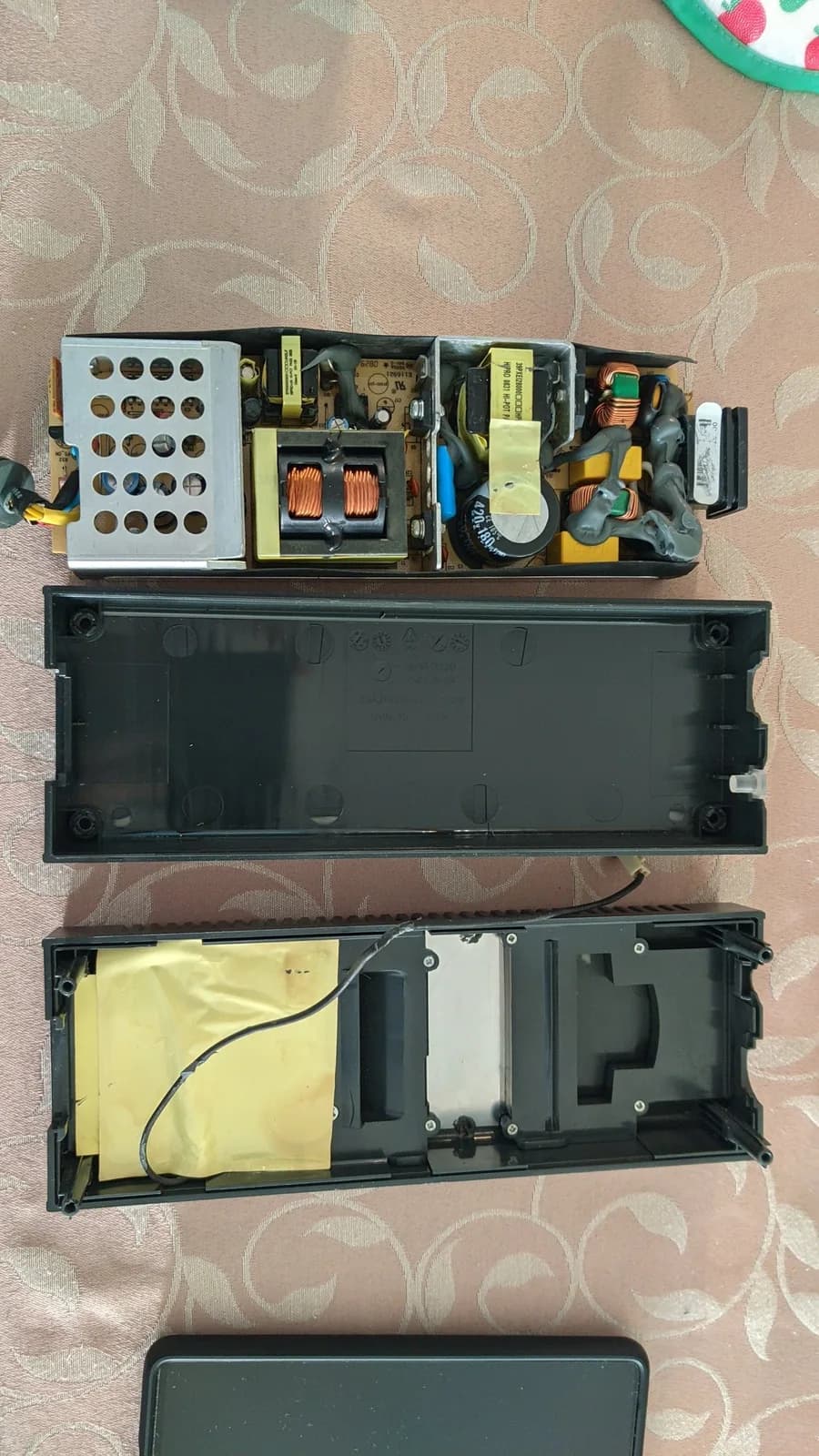 Xbox 360 S power supply fully disassembled on a patterned cloth: at top the single circuit board with its capacitors, transformers and a perforated metal shield, and below it the two empty halves of the black plastic case.