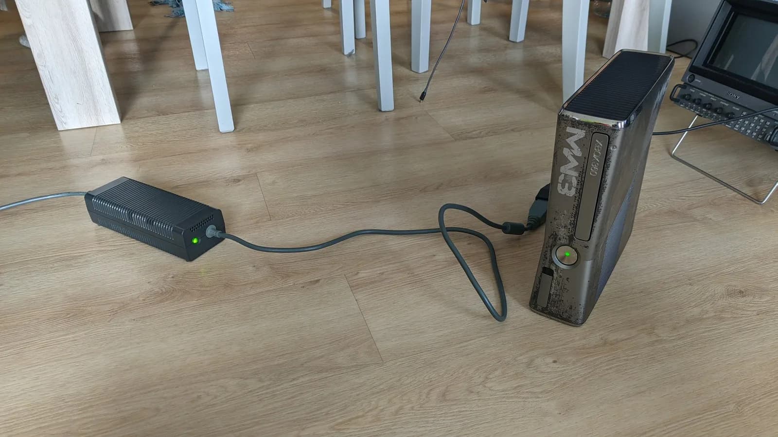 Xbox 360 S (Modern Warfare 3 special edition) standing upright on a wooden floor, powered on with its green LED lit, fed from its external power brick by the grey cable during post-repair testing, with a Sony BVM-D9H5J CRT monitor at the right edge.
